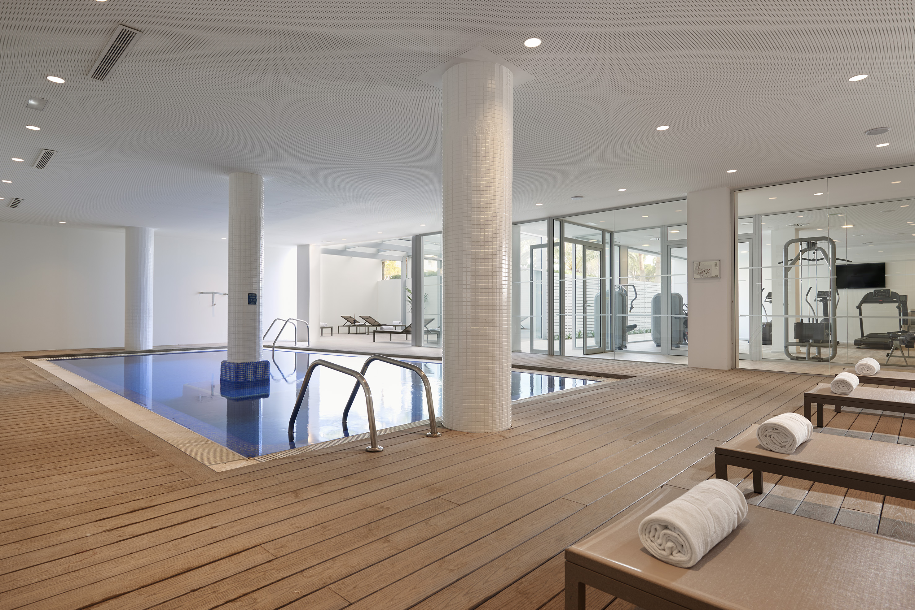 Bright indoor pool with wooden deck, lounge chairs, and adjacent fitness center.
