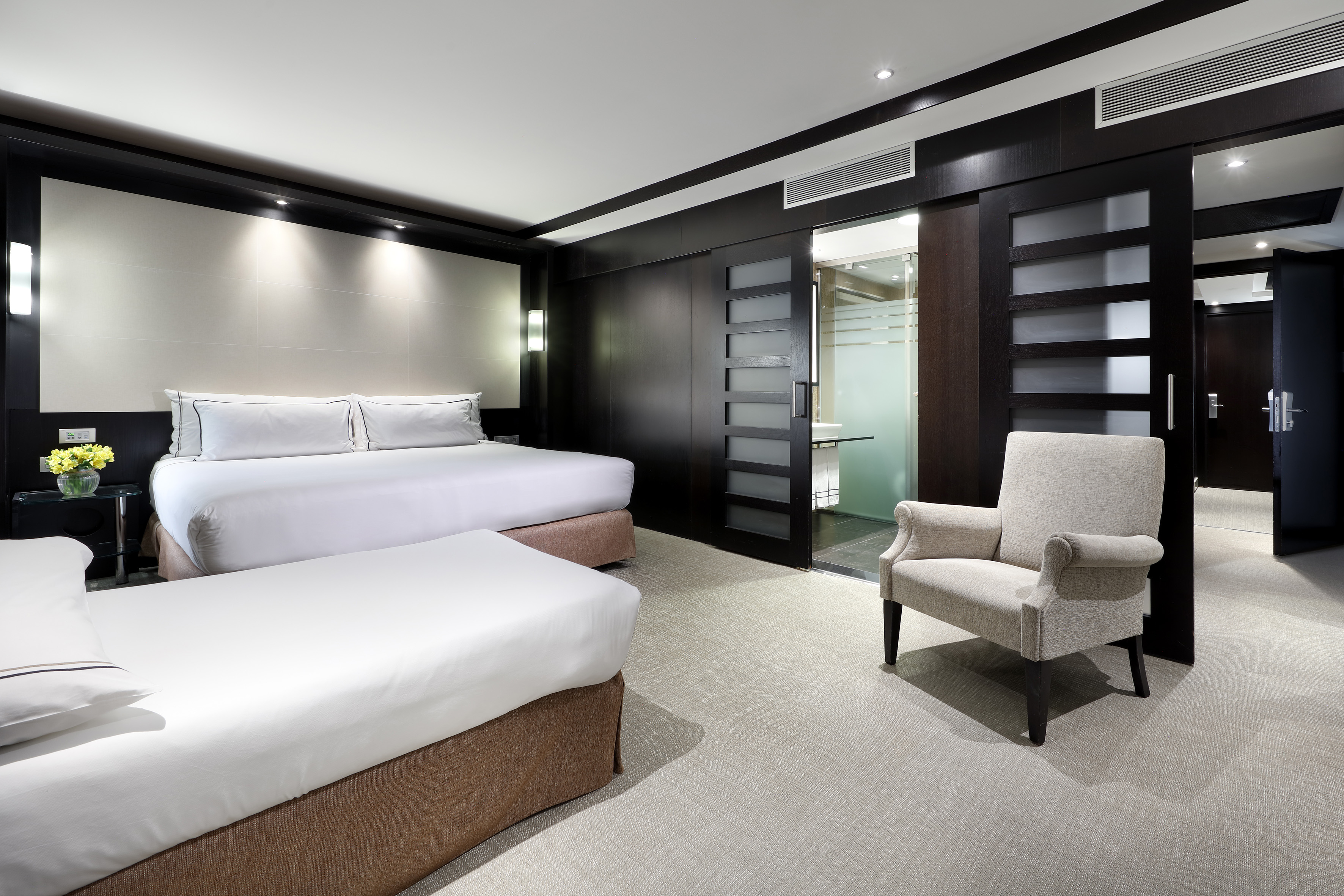 Modern hotel room with two beds, dark wood walls, armchair, and bathroom entrance.