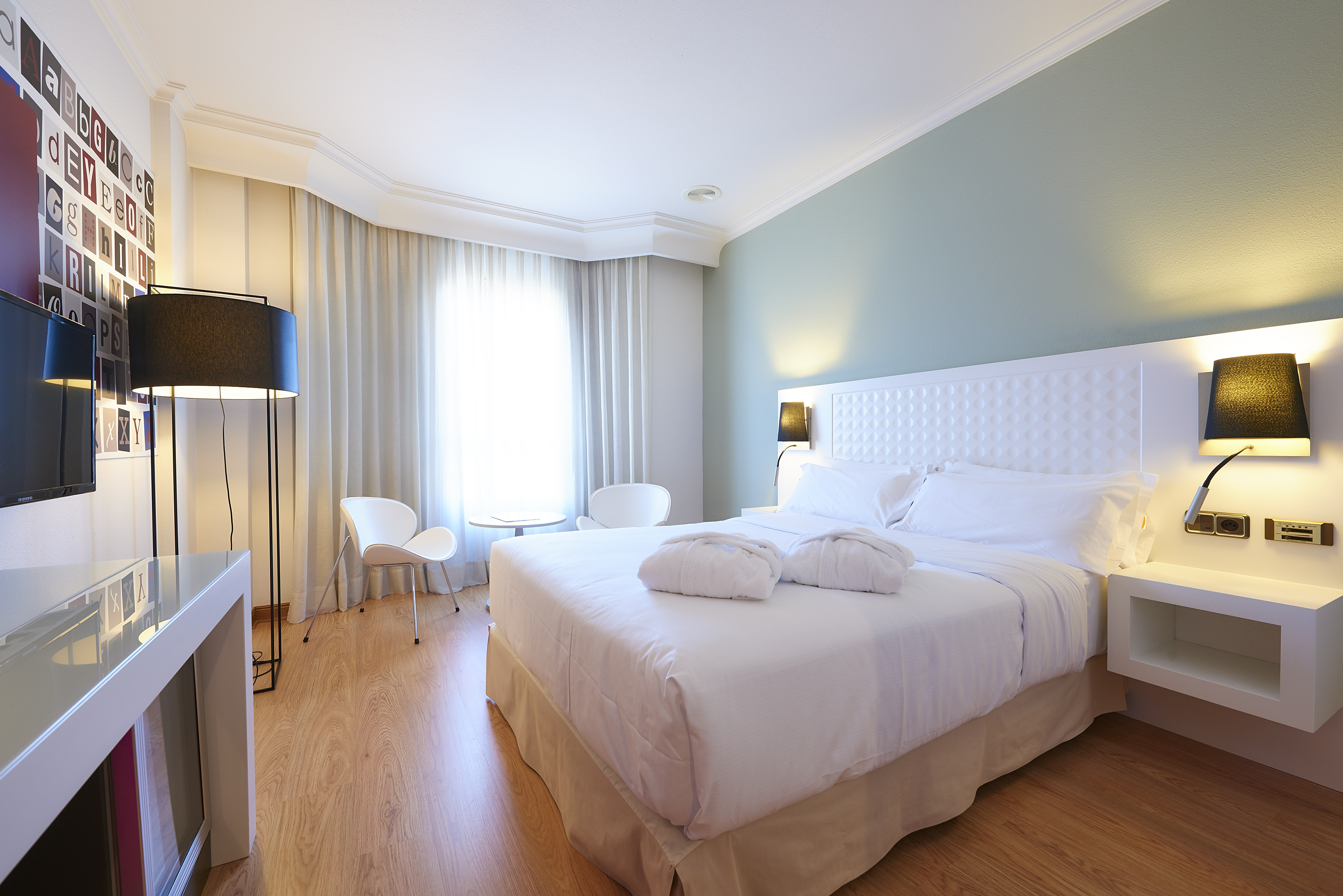 Modern hotel bedroom with white bed, bathrobes, light green wall, and wood floor.