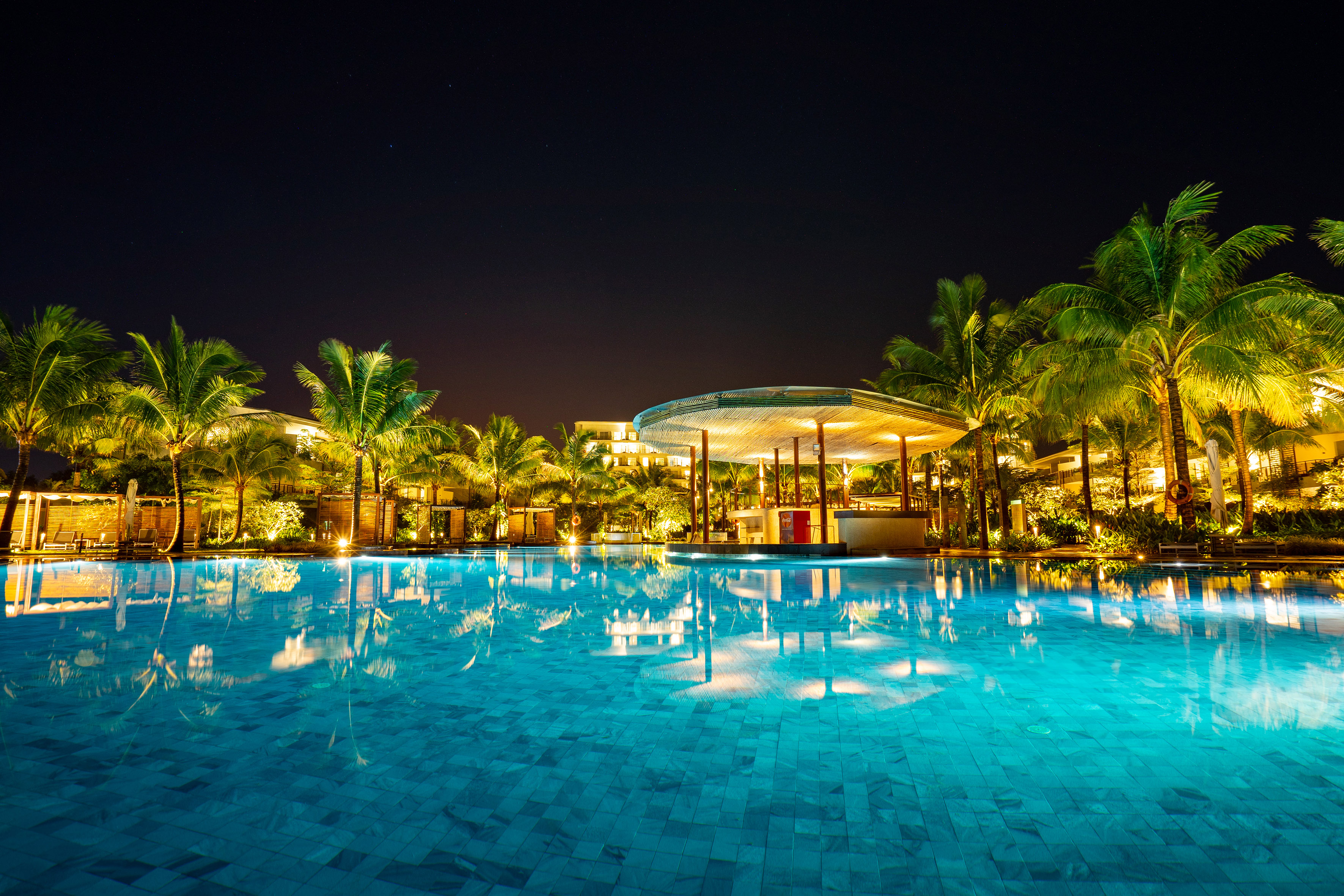 Luxury resort swimming pool at night with illuminated palm trees and bar.