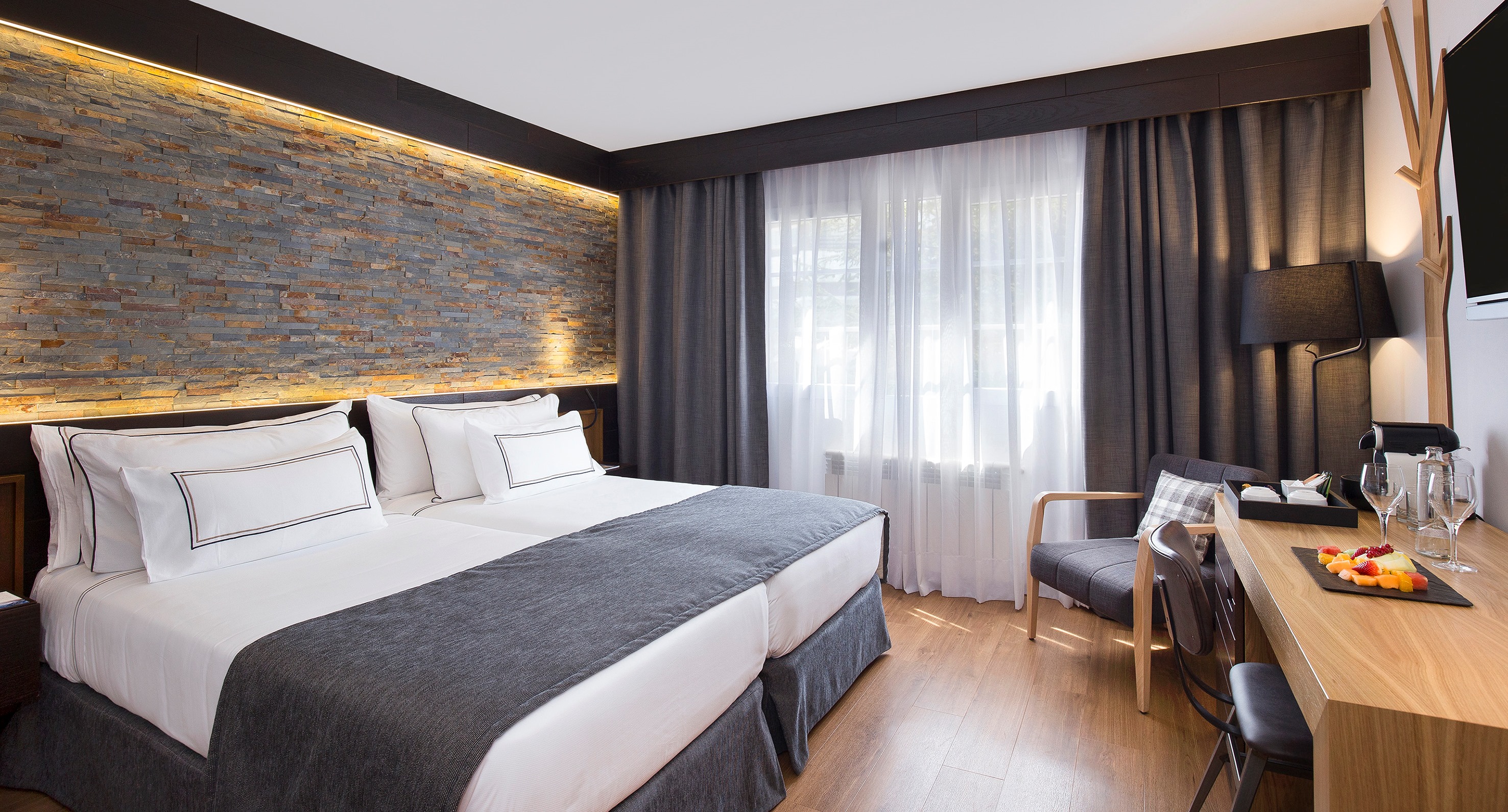 Modern hotel room with twin beds, stone accent wall, desk, and armchair.