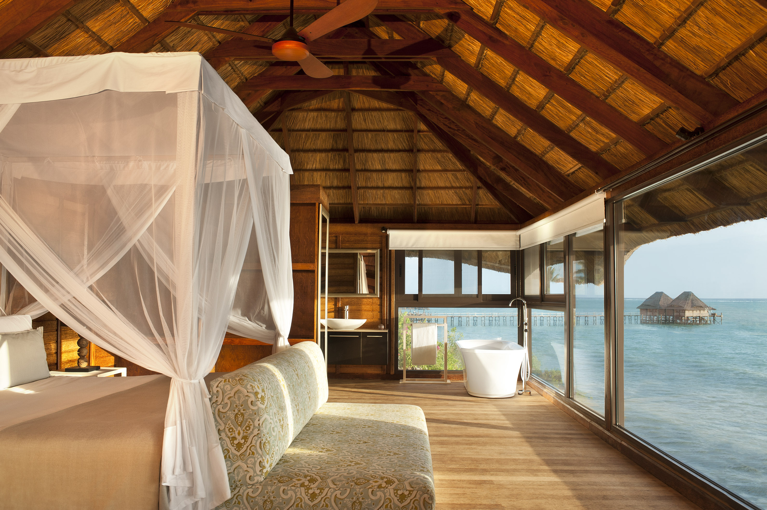 Tropical overwater bungalow interior: canopy bed, ocean view, freestanding tub.