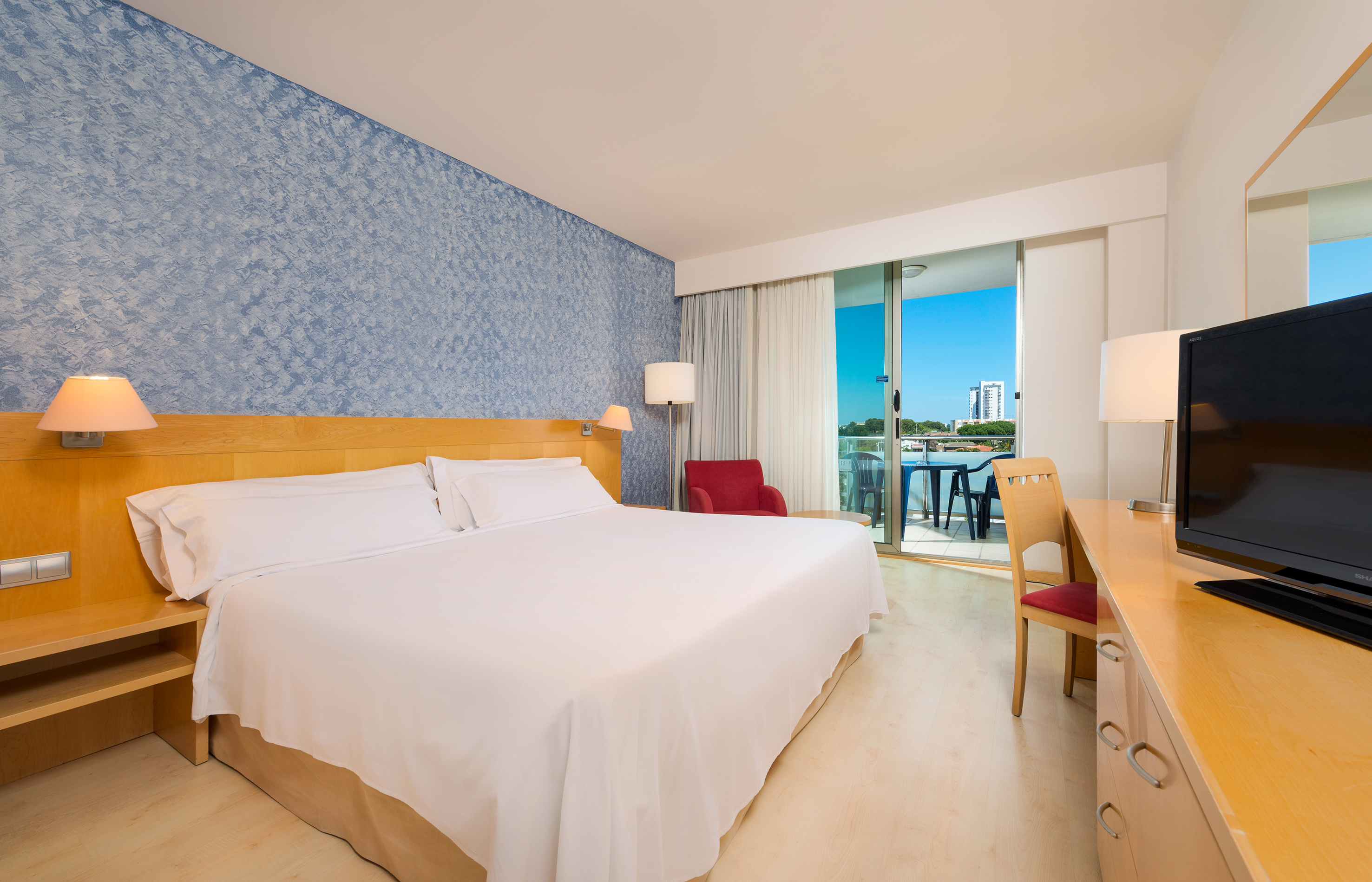 Hotel room with white bed, blue wall, wooden furniture, and sunny balcony.