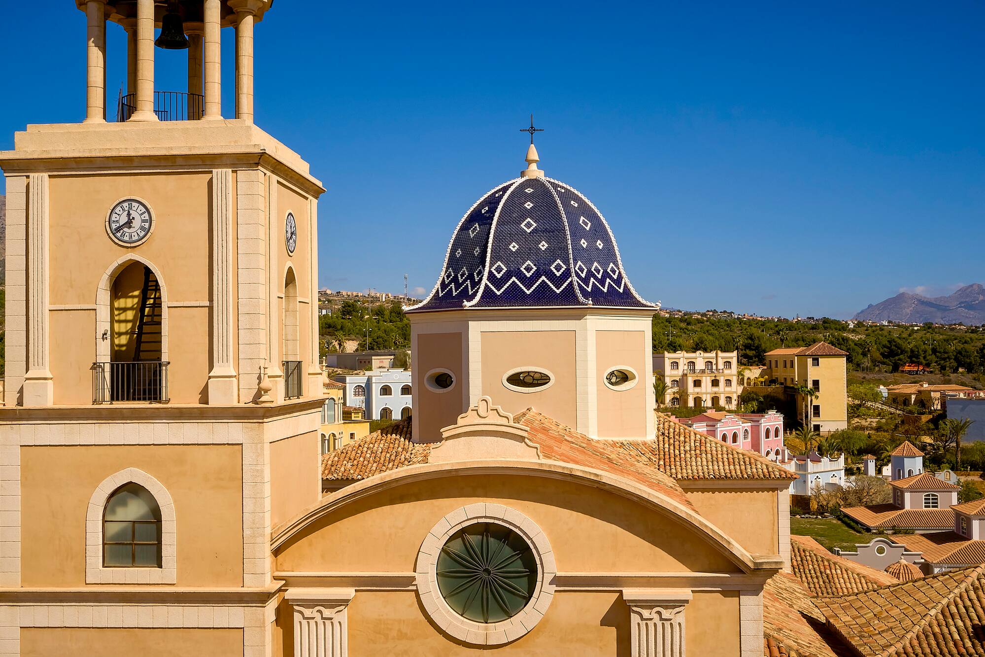 Beige church with blue patterned dome, clock tower, and terracotta roofs in sunny town.