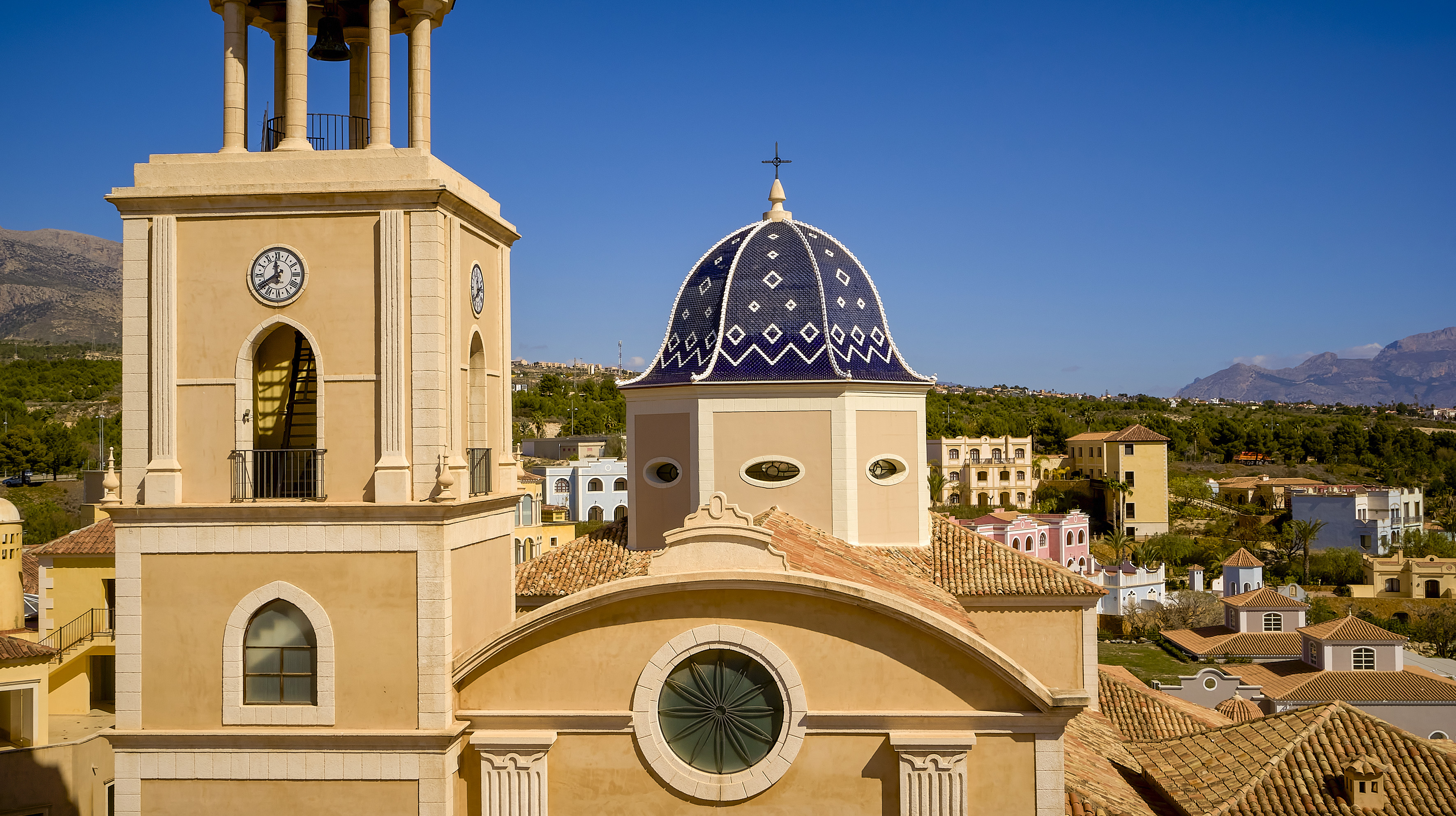 Beige church with blue patterned dome, clock tower, and terracotta roofs in sunny town.