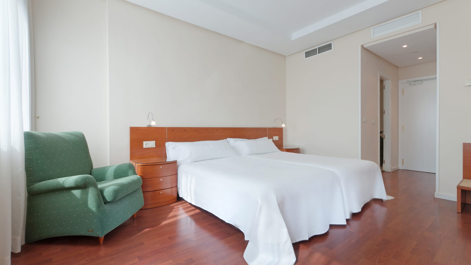 Hotel guest room with two beds, green armchair, and polished hardwood floor.