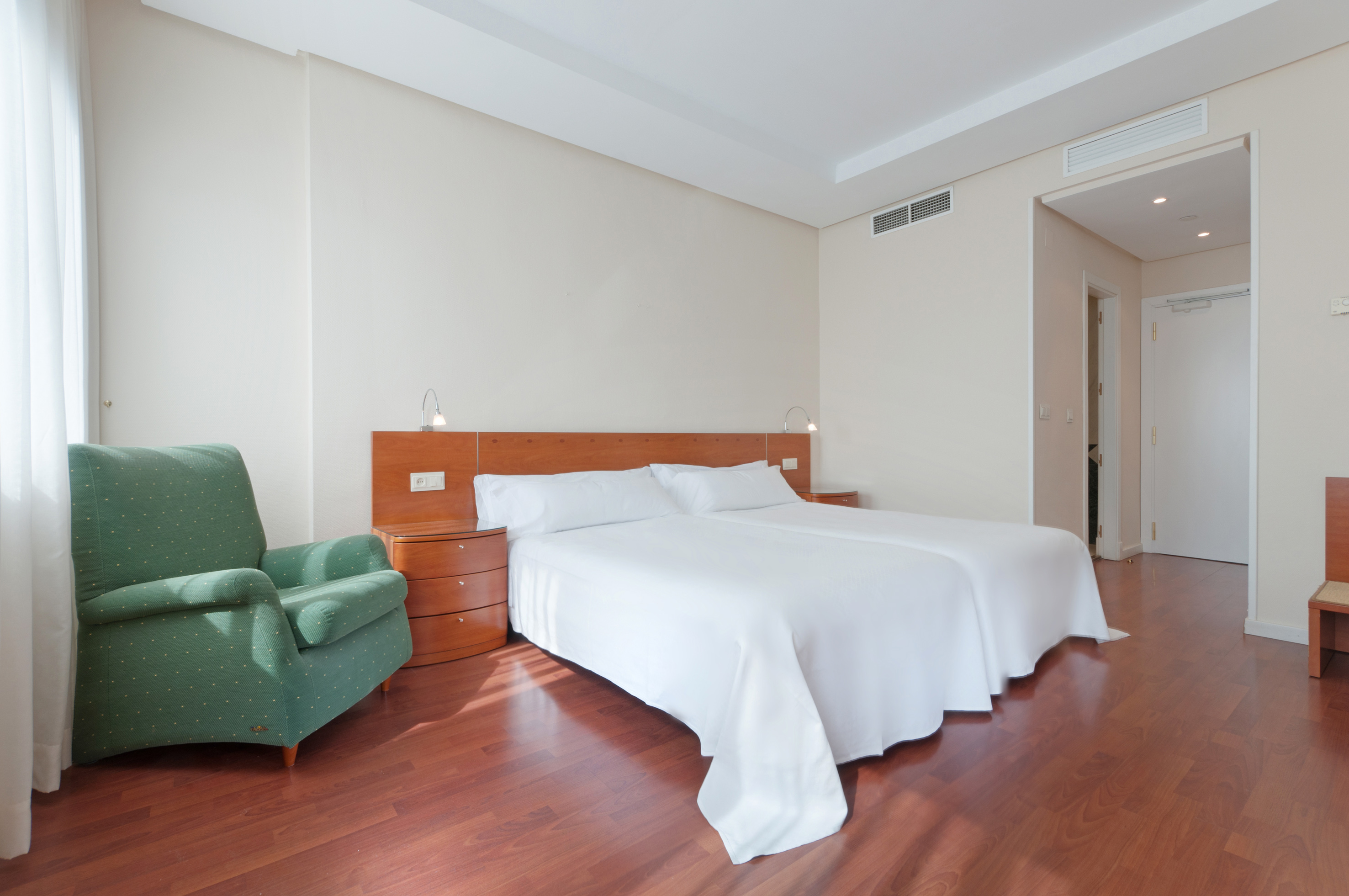 Hotel guest room with two beds, green armchair, and polished hardwood floor.