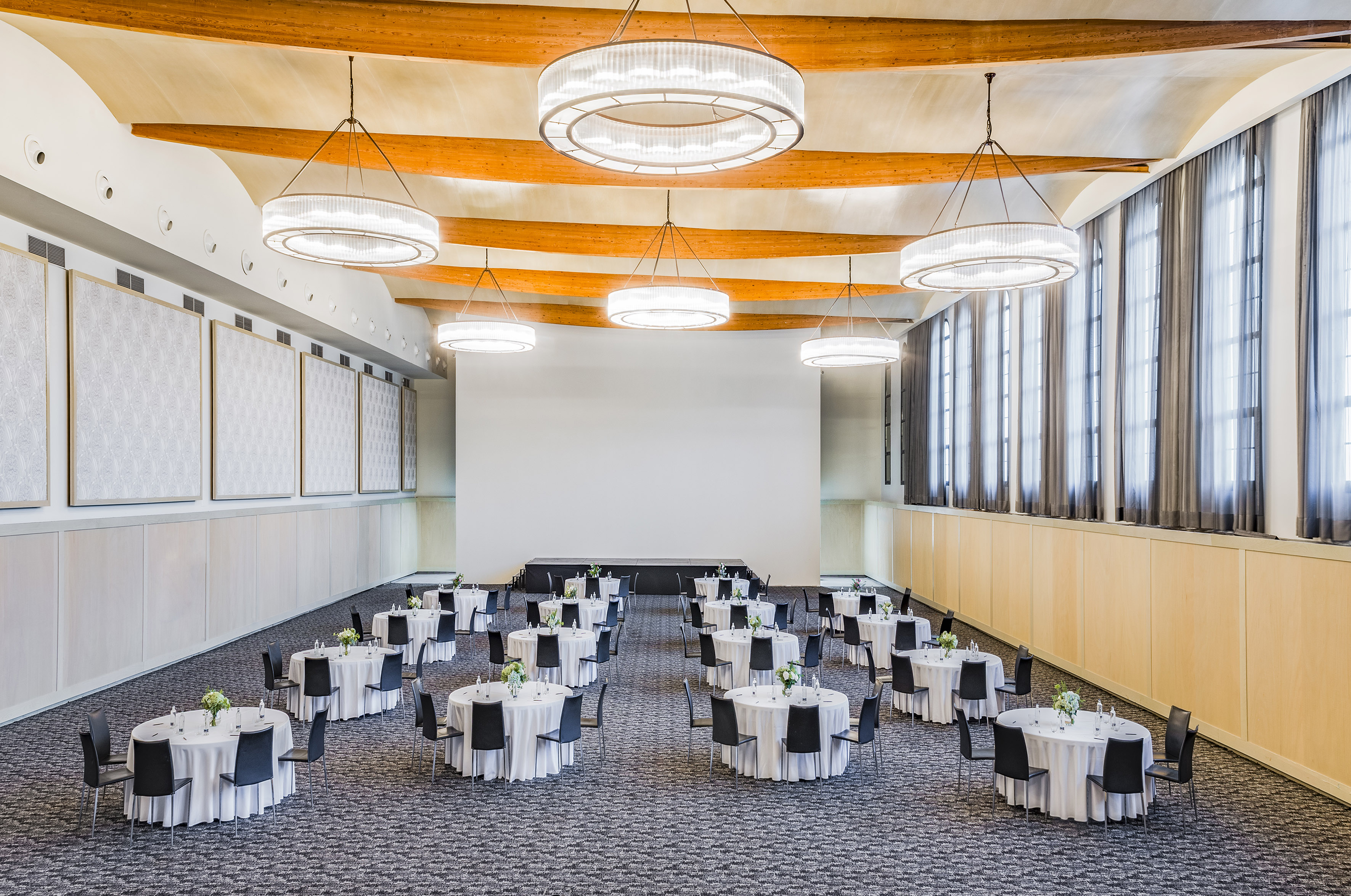 Conference ballroom with round tables, chairs, stage, and modern chandeliers.