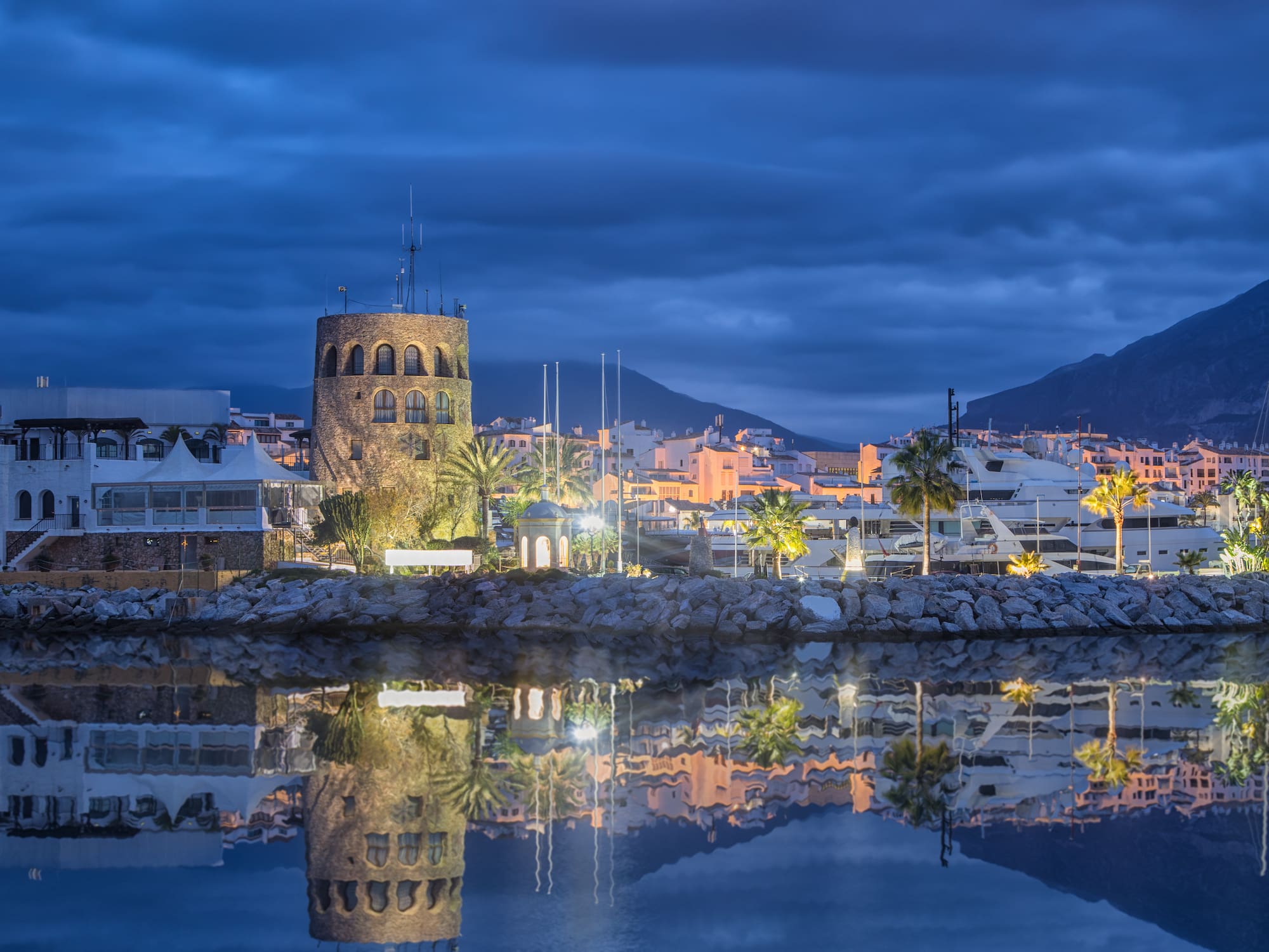 Dusk marina with illuminated stone tower, yachts, and water reflections.