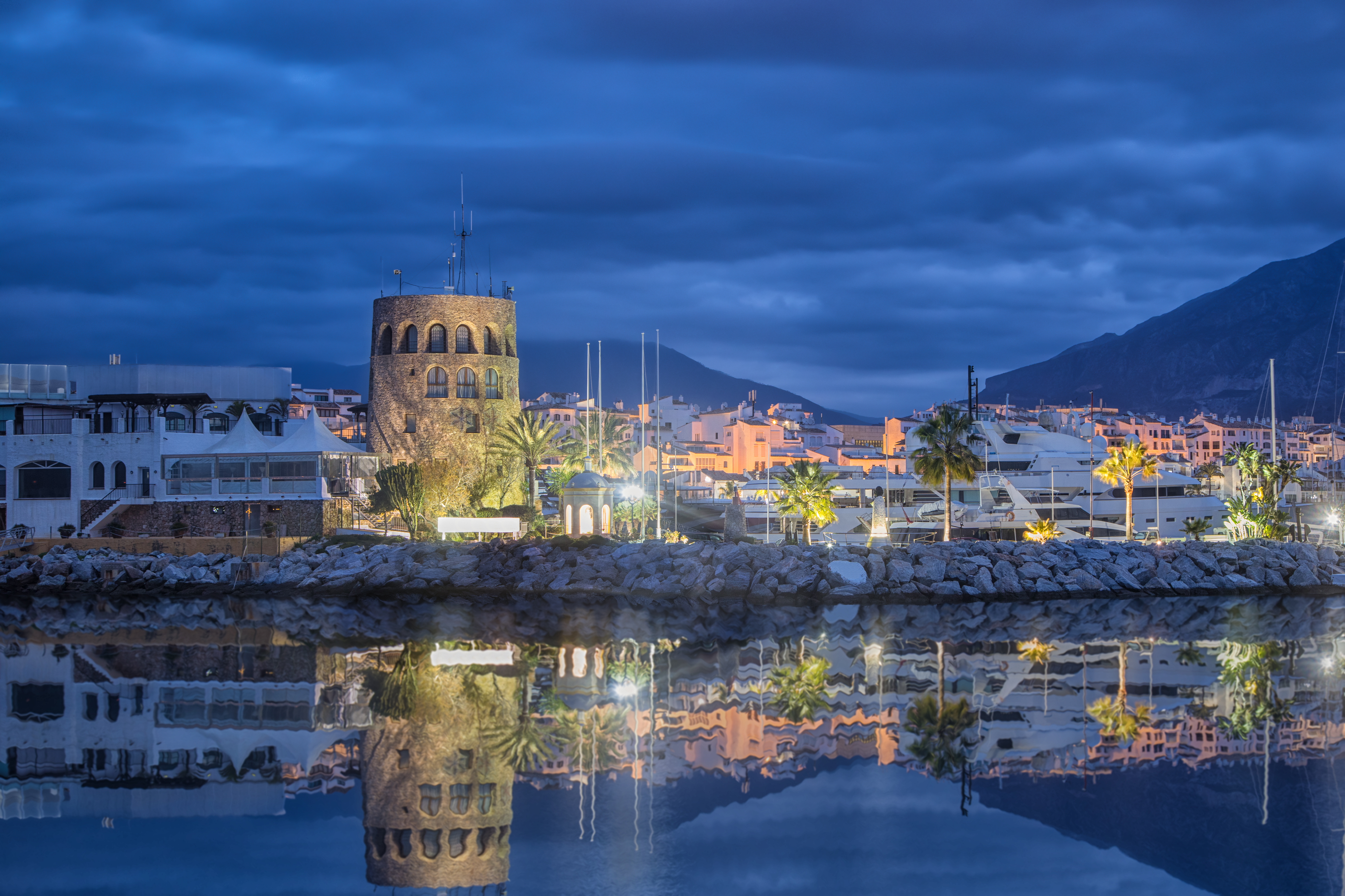 Dusk marina with illuminated stone tower, yachts, and water reflections.