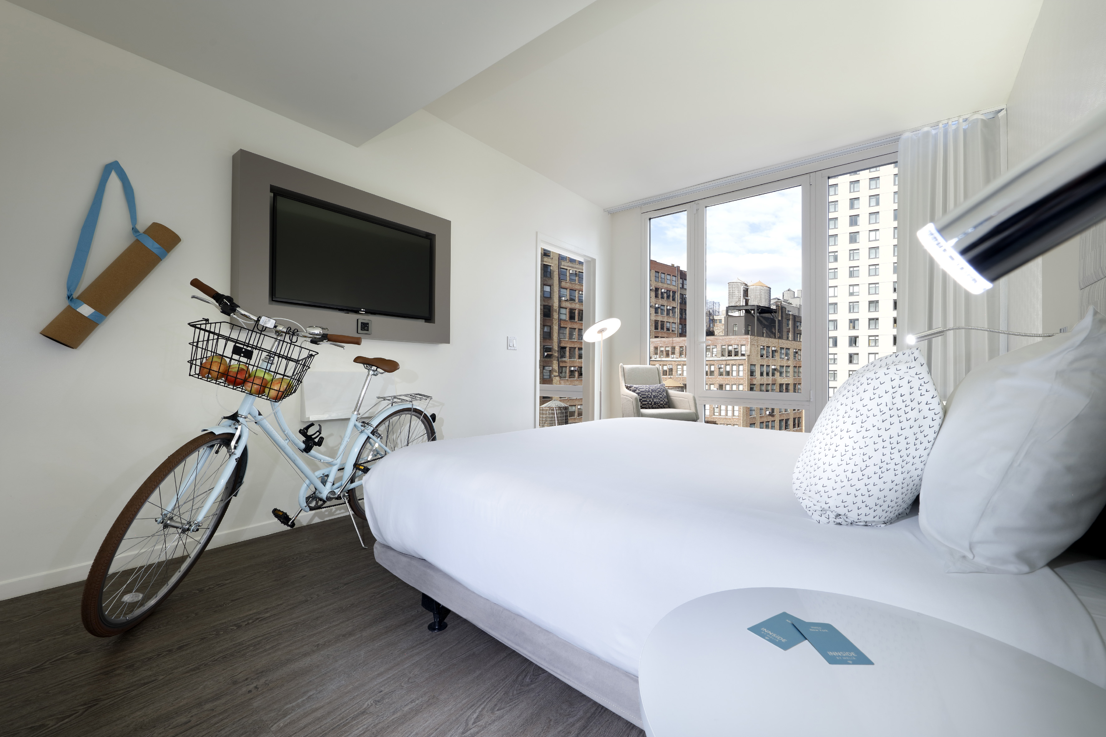 Modern hotel room with city view, bed, bicycle, yoga mat, and wall TV.