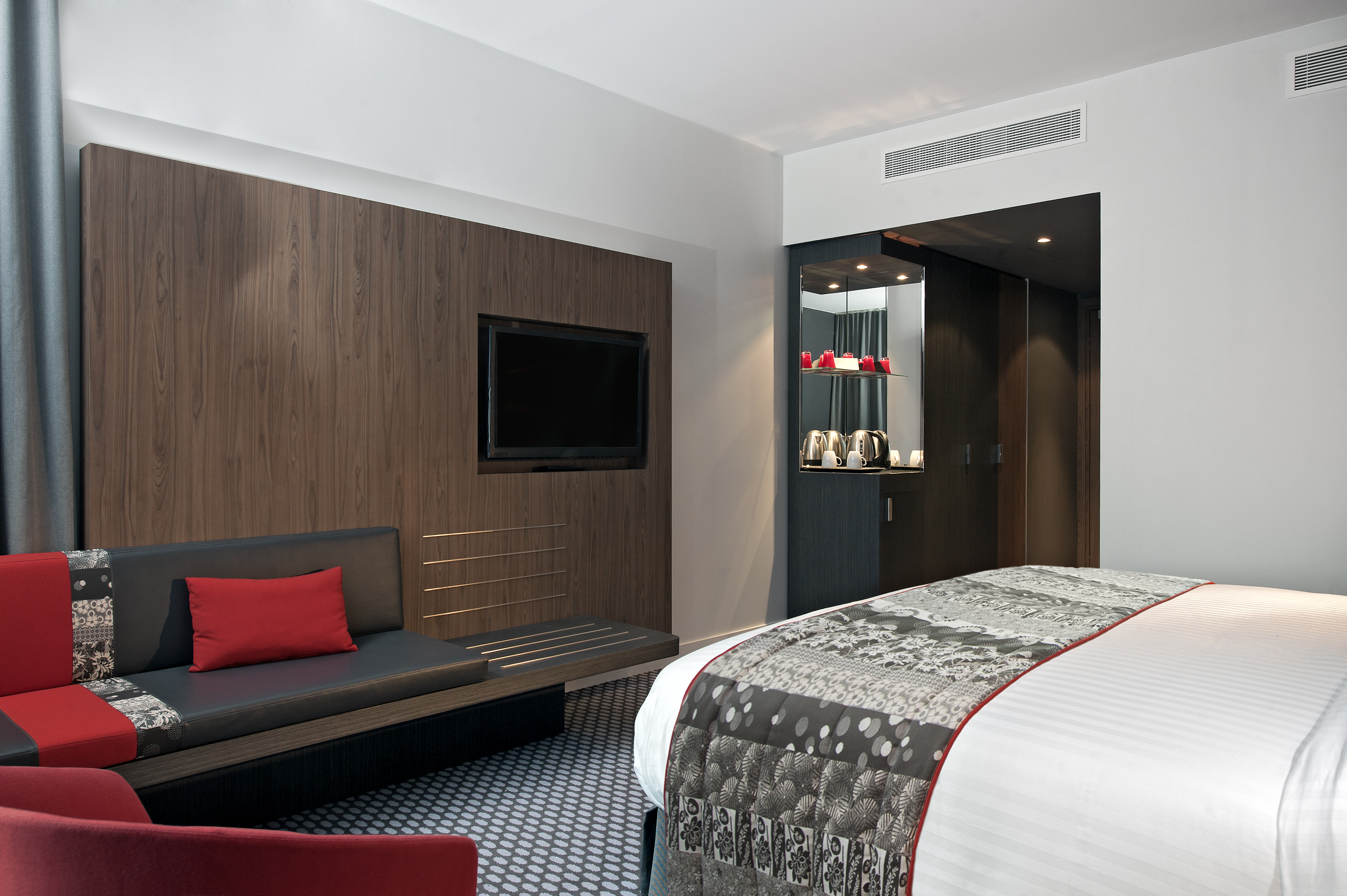 Sleek hotel room with bed, TV on wood panel, red sofa, and mini-bar.