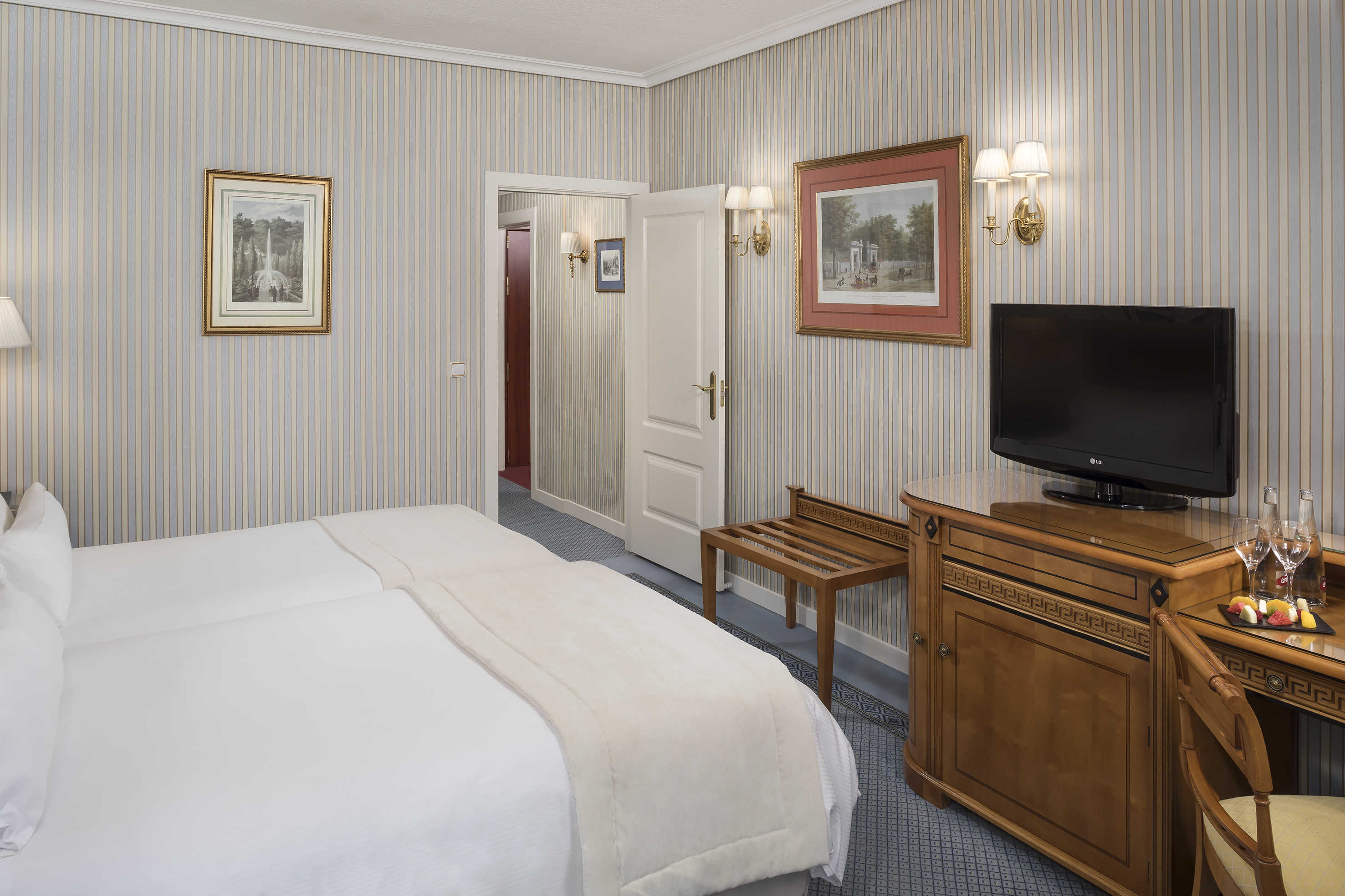 Hotel room with twin beds, striped wallpaper, wooden furniture, TV, and desk.