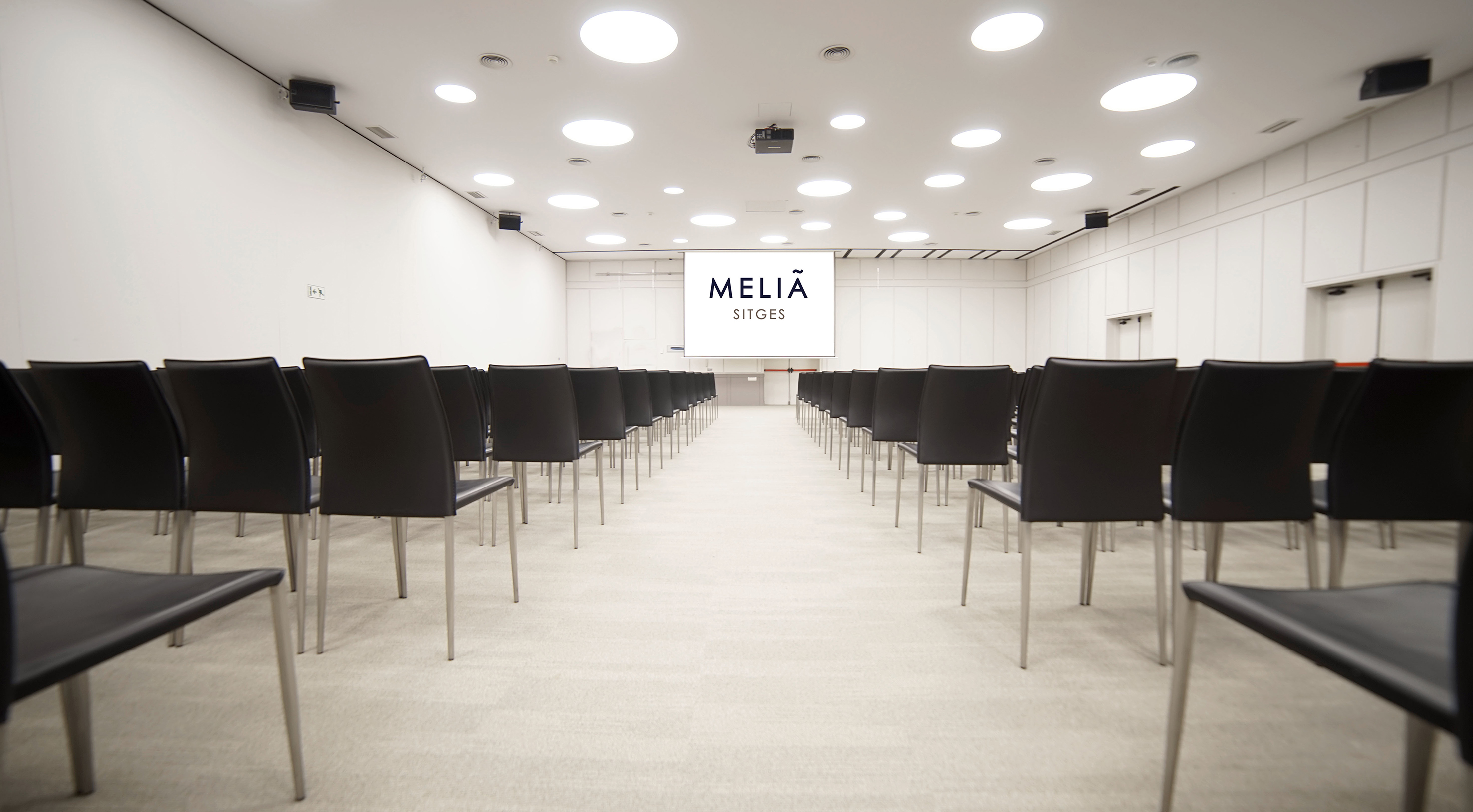 Empty Meliã Sitges conference room with rows of black chairs and projector screen.