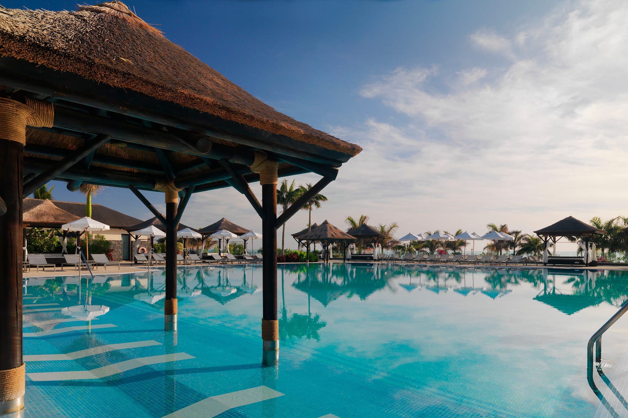 Tropical resort pool with thatched cabana, sun loungers, palm trees.