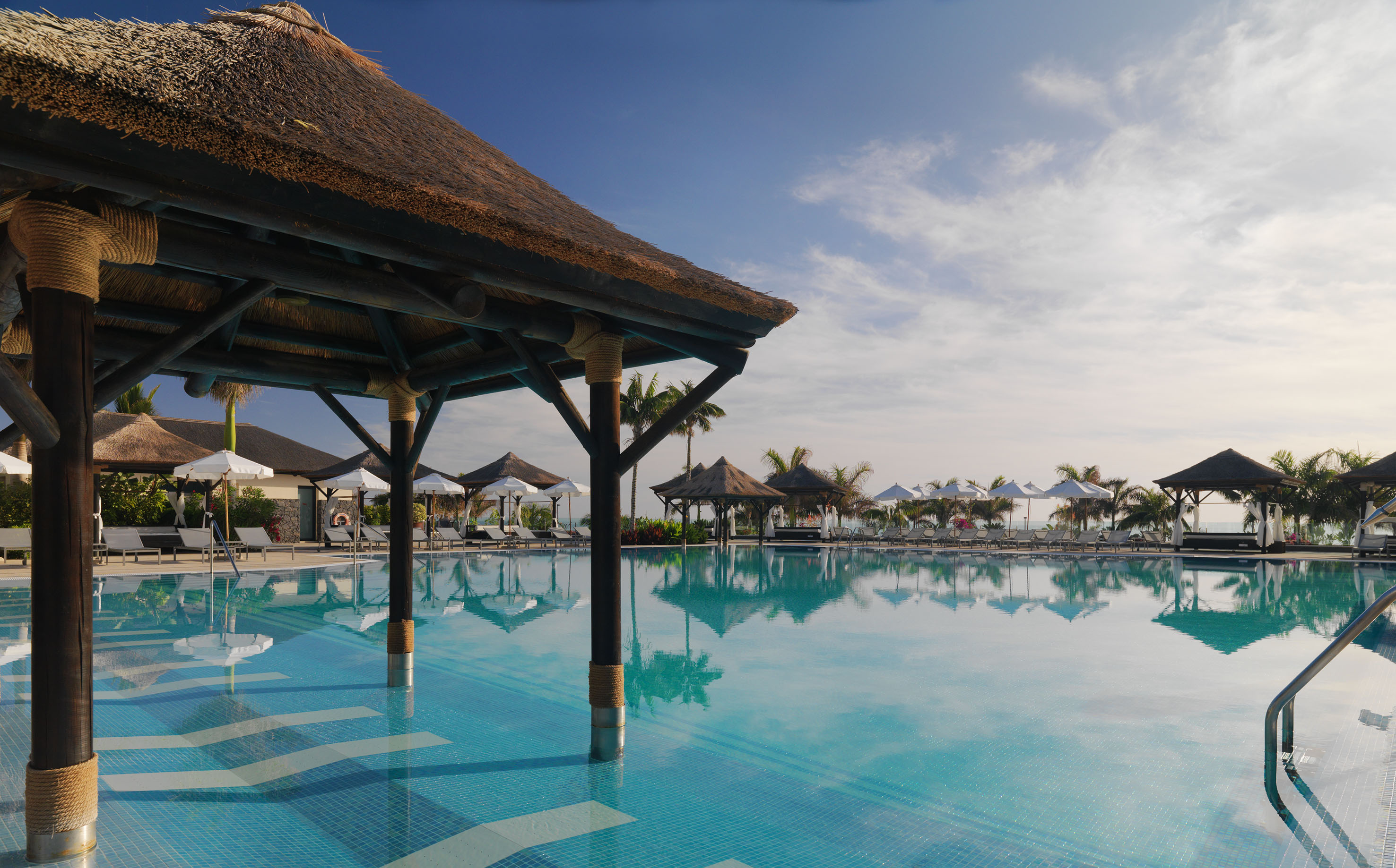 Tropical resort pool with thatched cabana, sun loungers, palm trees.