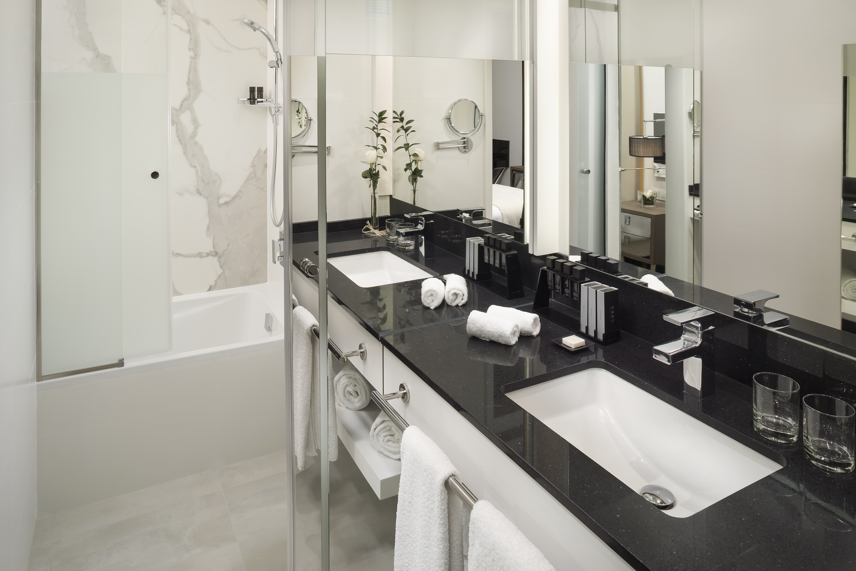 Modern hotel bathroom: black double vanity, white sinks, shower, marble details.