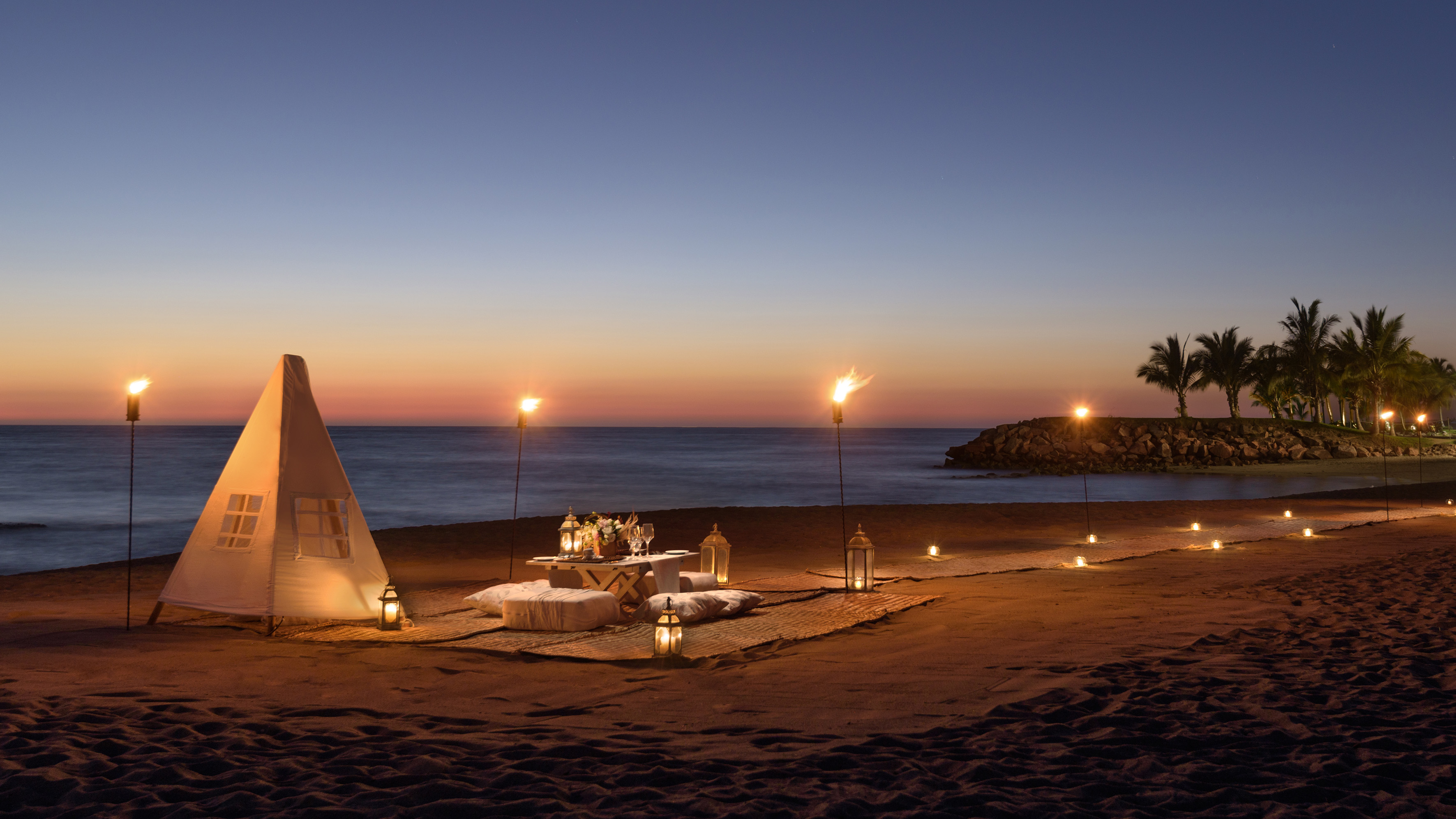 Romantic beach dinner with tent, tiki torches, and lanterns at sunset.