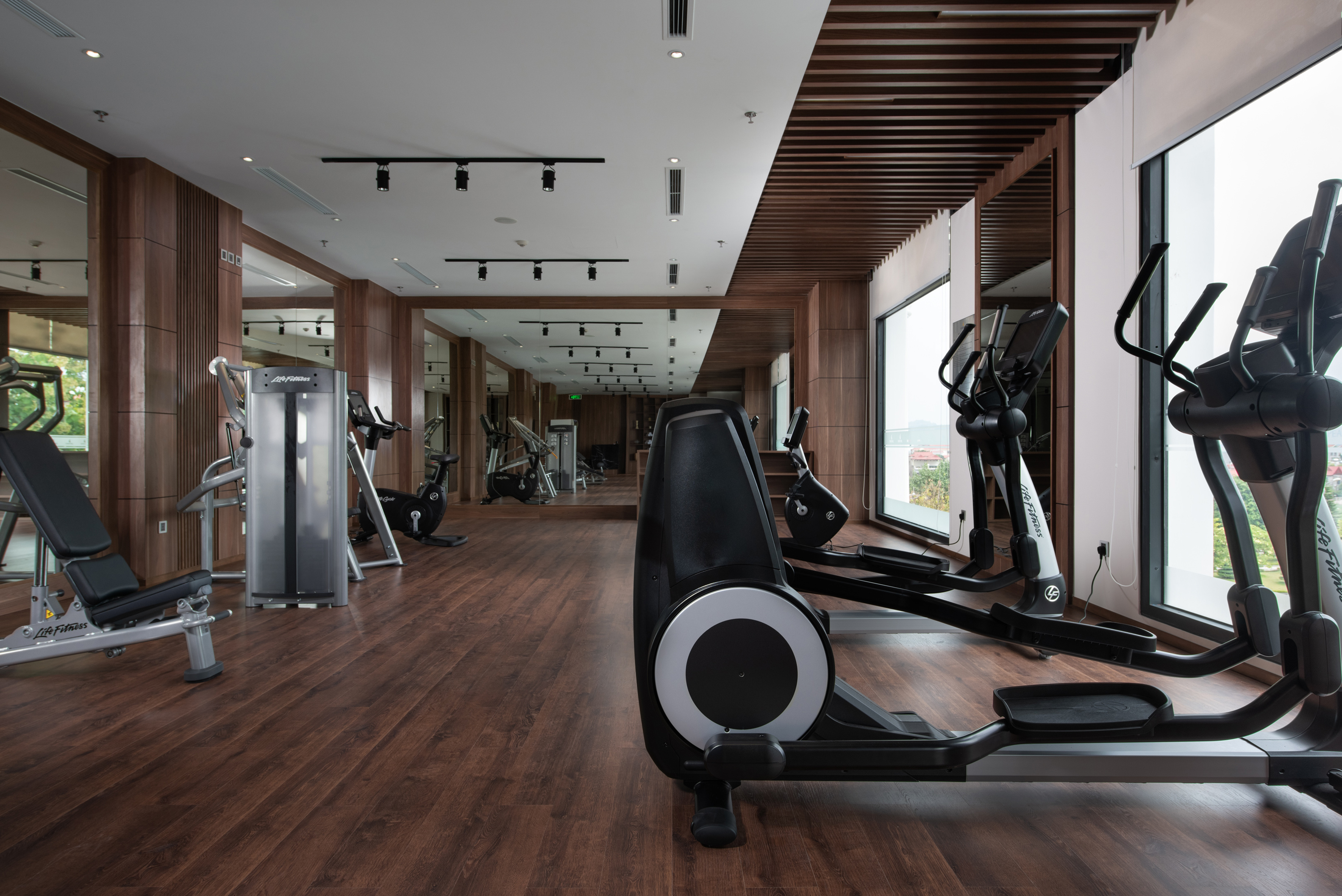 Modern gym with ellipticals, weight bench, cardio equipment, wood floor.