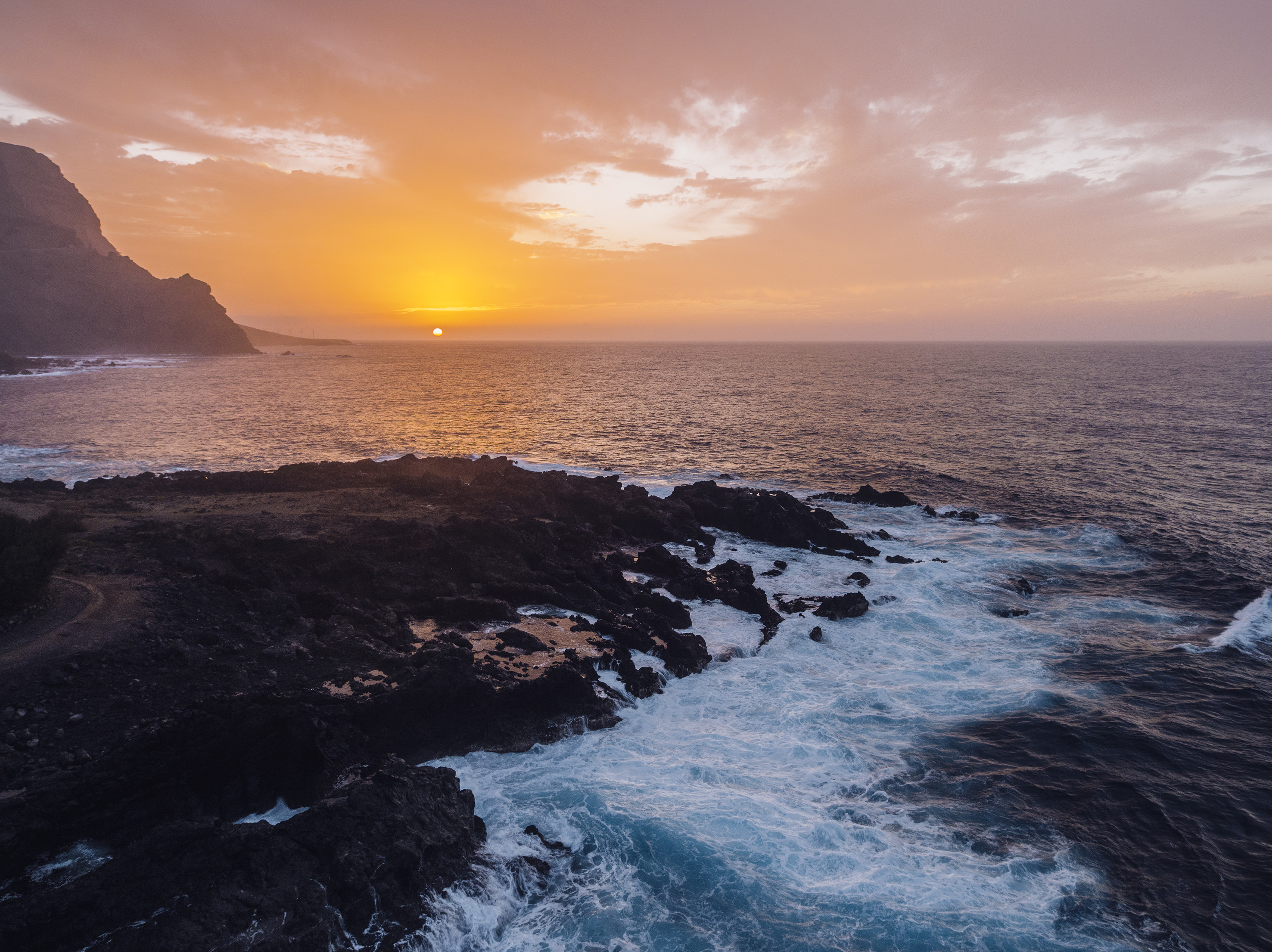 Dramatic sunset over dark rocky coast, ocean waves, and distant cliffs.