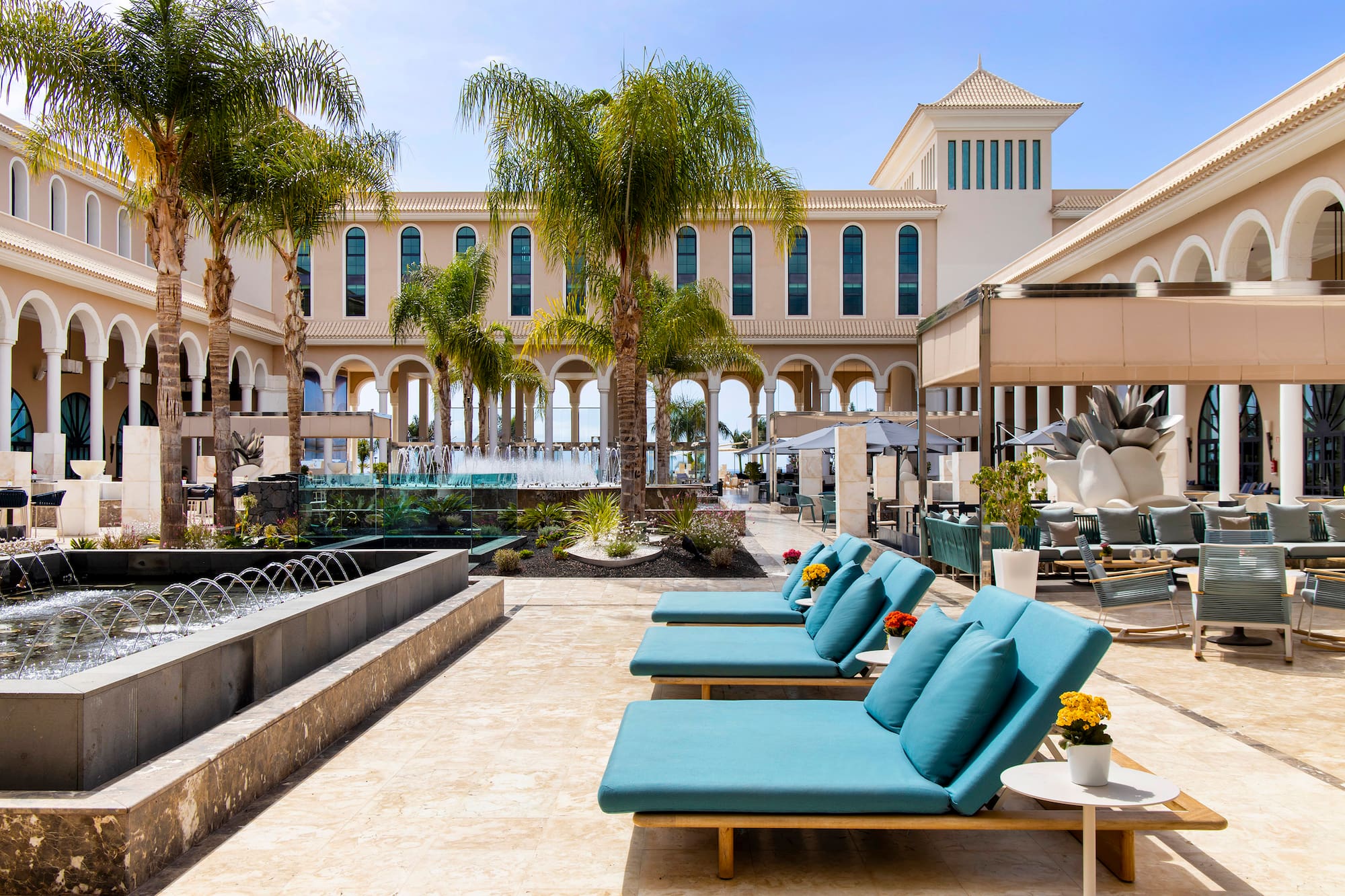 Luxury resort courtyard with palm trees, fountains, and blue sun loungers.