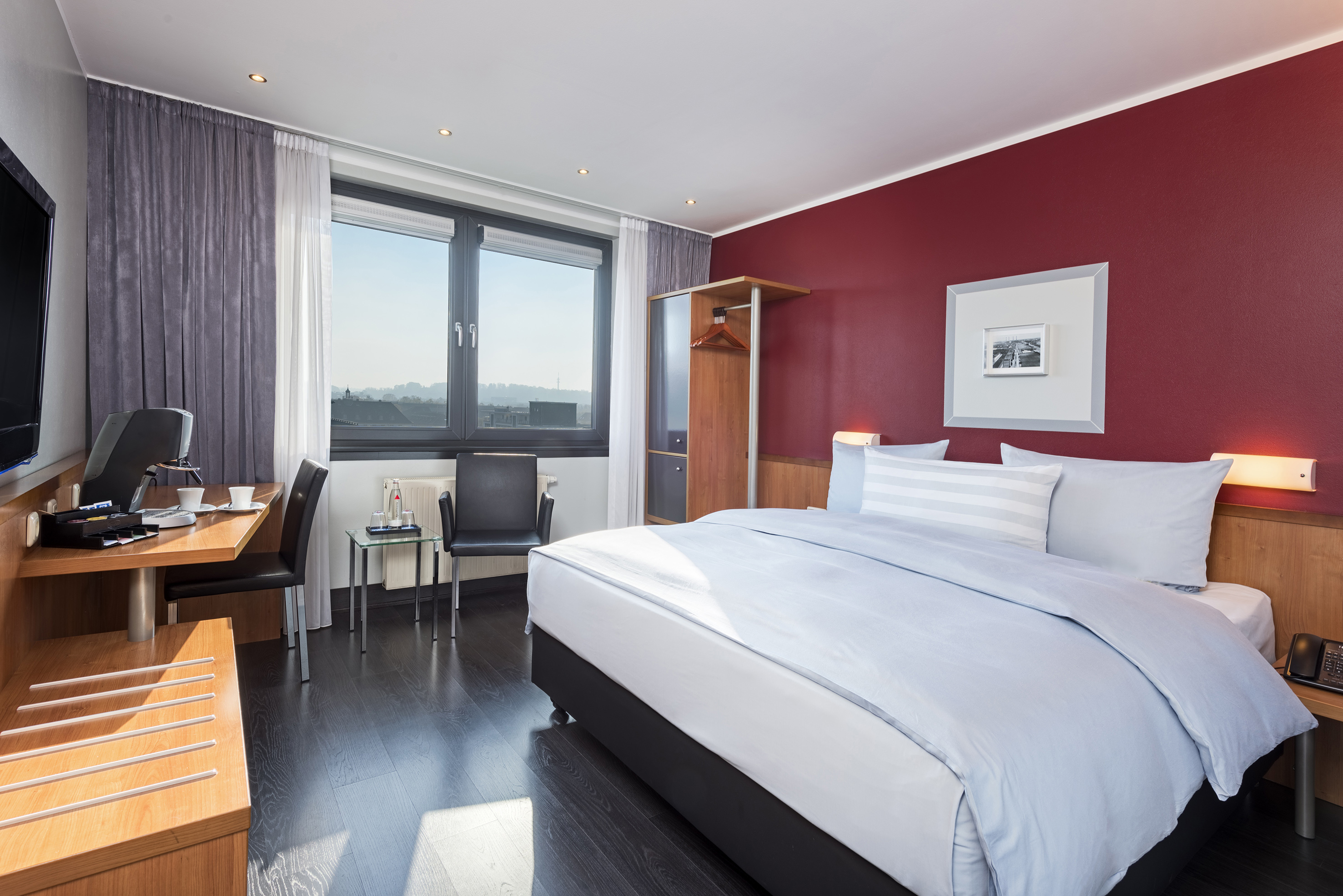 Modern hotel room with king bed, red accent wall, desk, and city view.