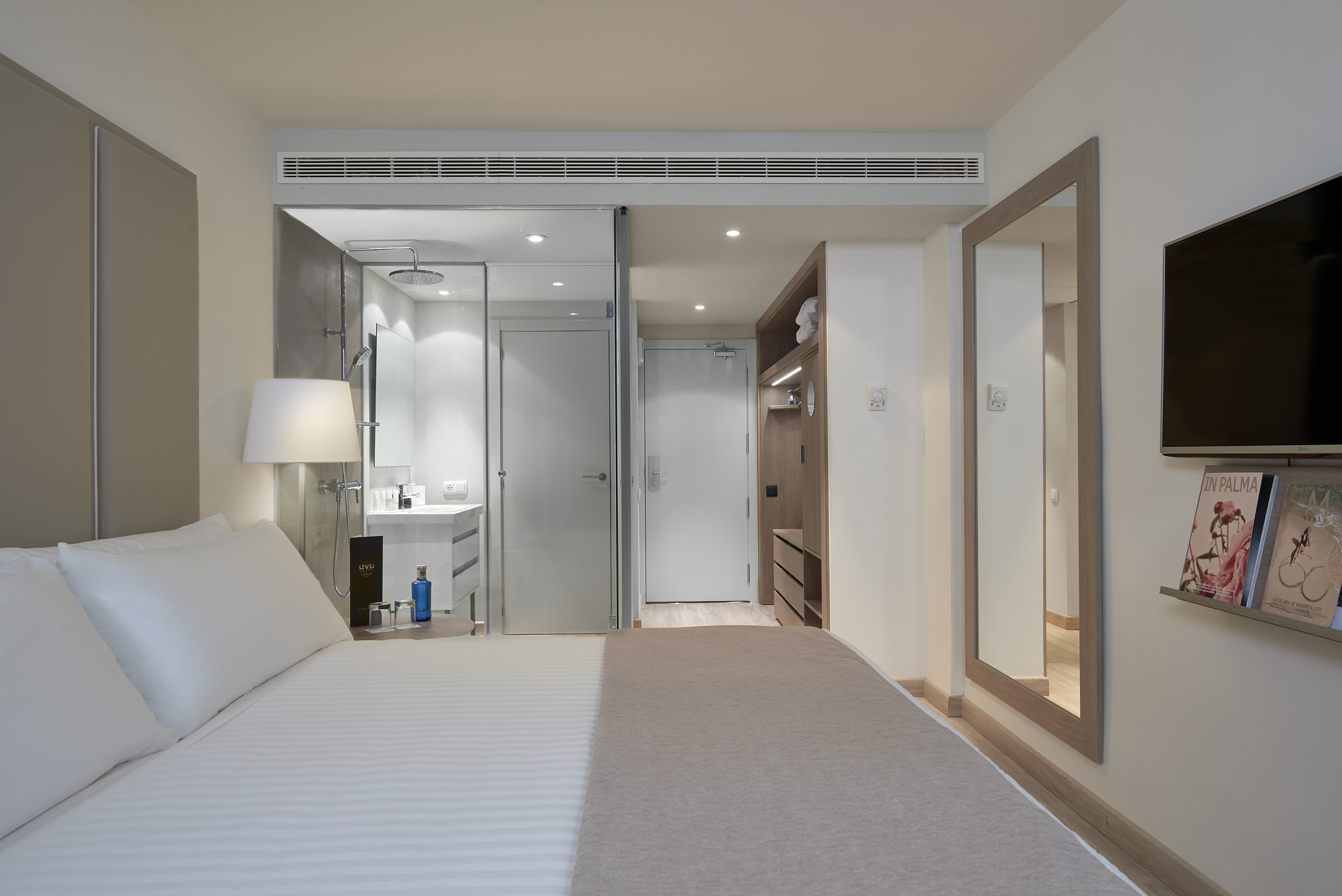 Modern hotel guest room with bed, open shower, vanity, wall TV, and mirror.
