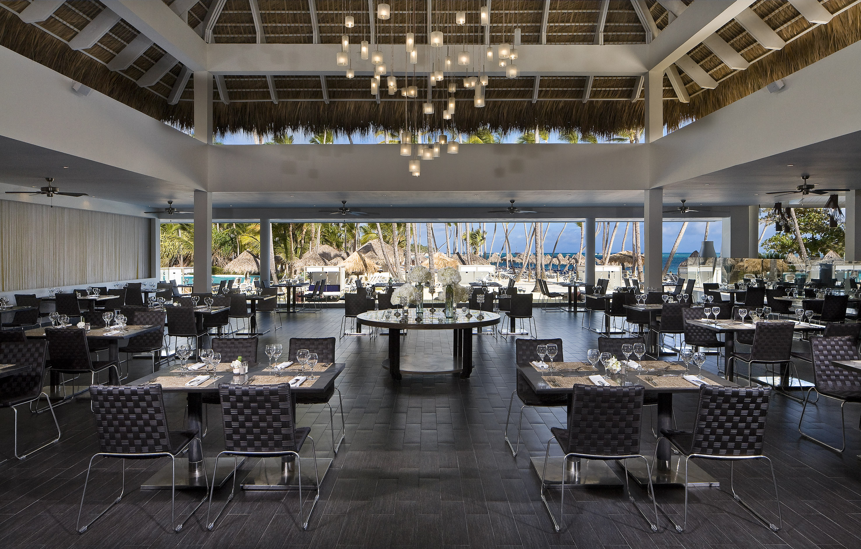 Tropical resort restaurant with ocean view, modern decor, and thatched roof.