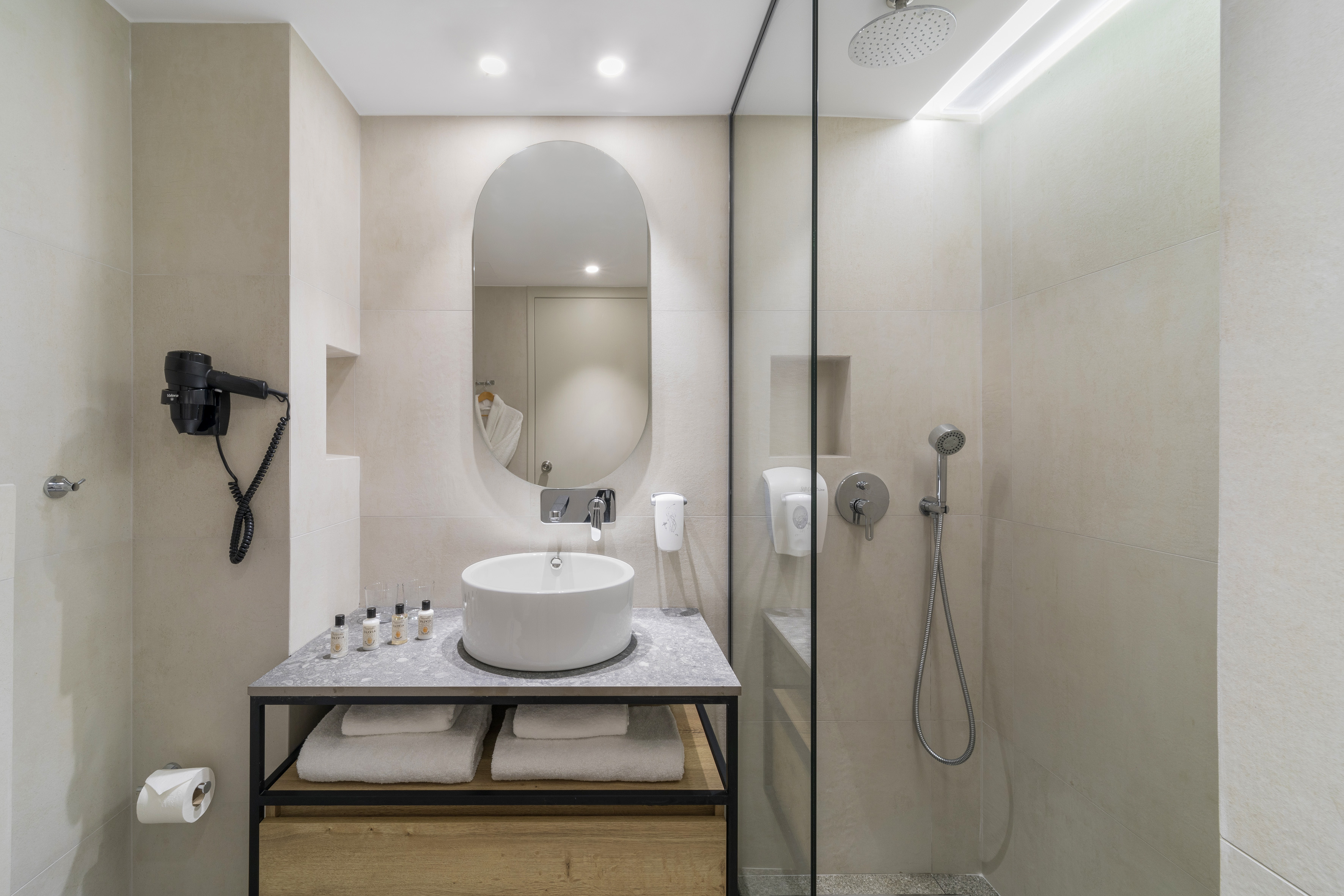Modern hotel bathroom with beige tiles, vessel sink, oval mirror, and glass shower.