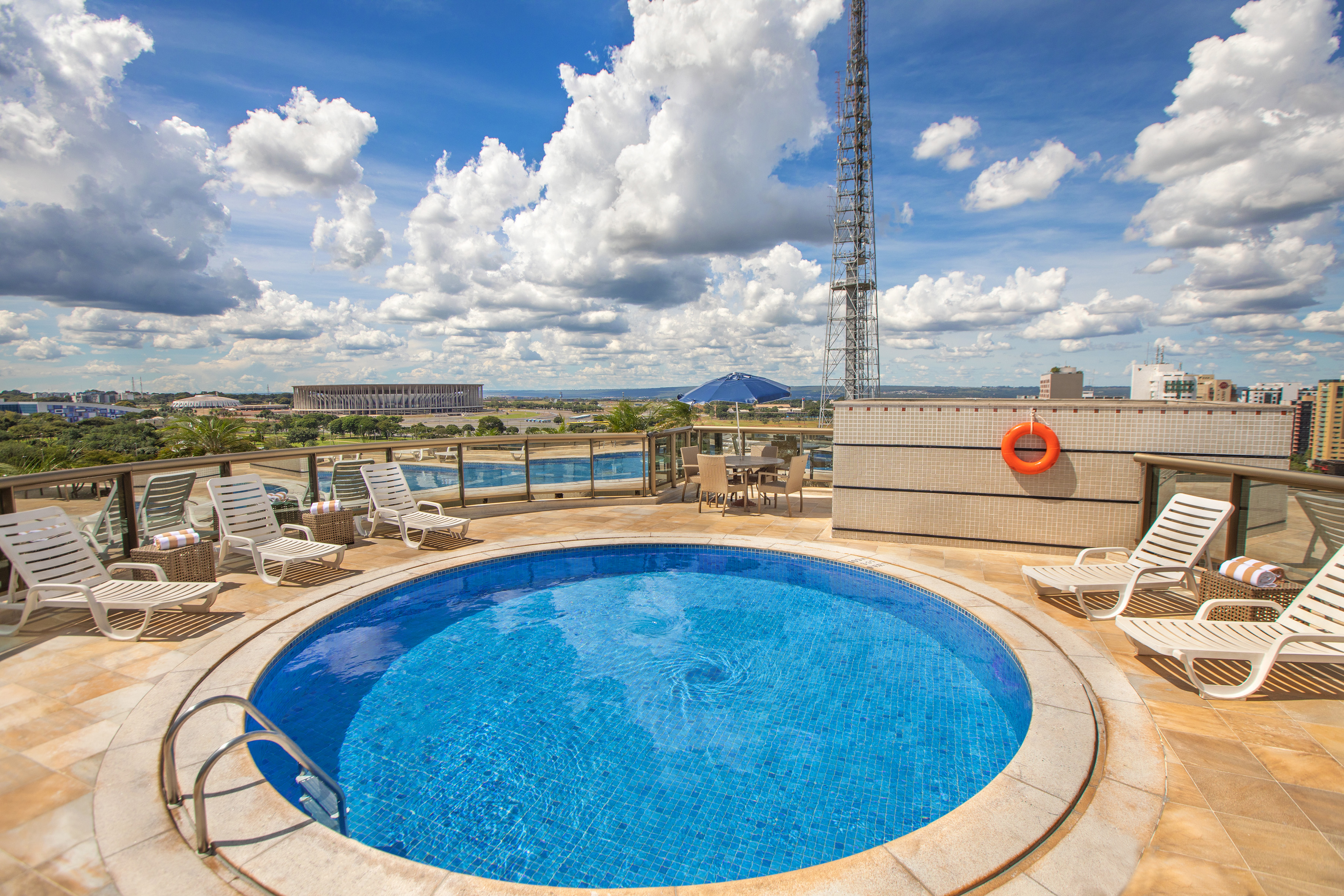 Rooftop pool, lounge chairs, city skyline with Brasília stadium under blue sky.