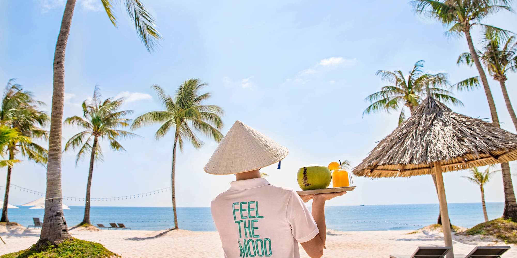 a person wearing a conical hat holding a tray with fruit and a straw umbrella on a beach