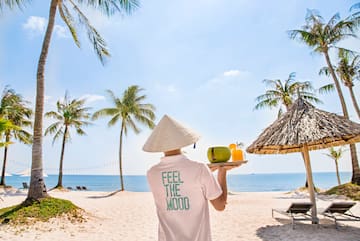 a person wearing a conical hat holding a tray with fruit and a straw umbrella on a beach