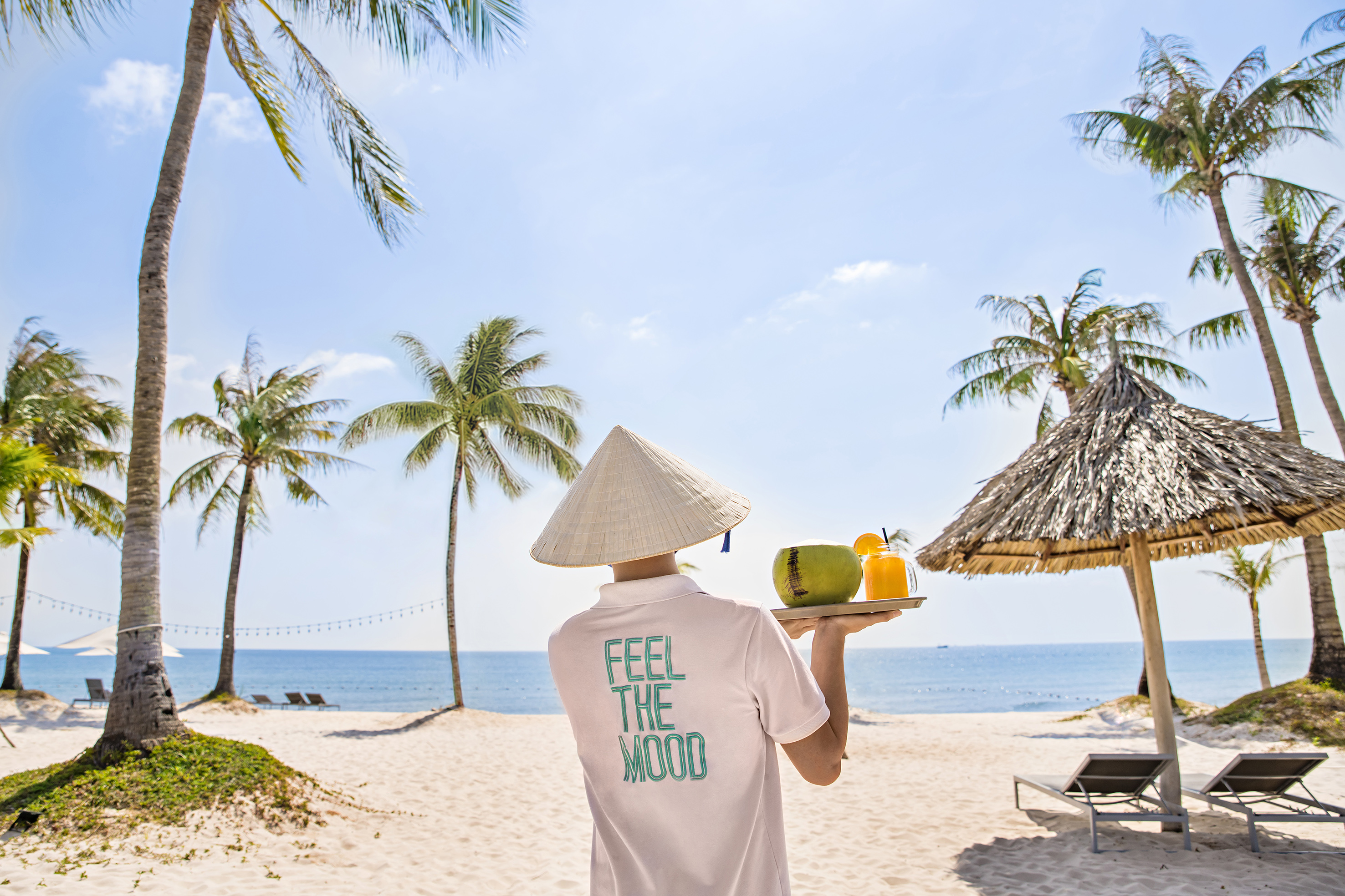 Person in conical hat on tropical beach holding fresh coconut and juice.