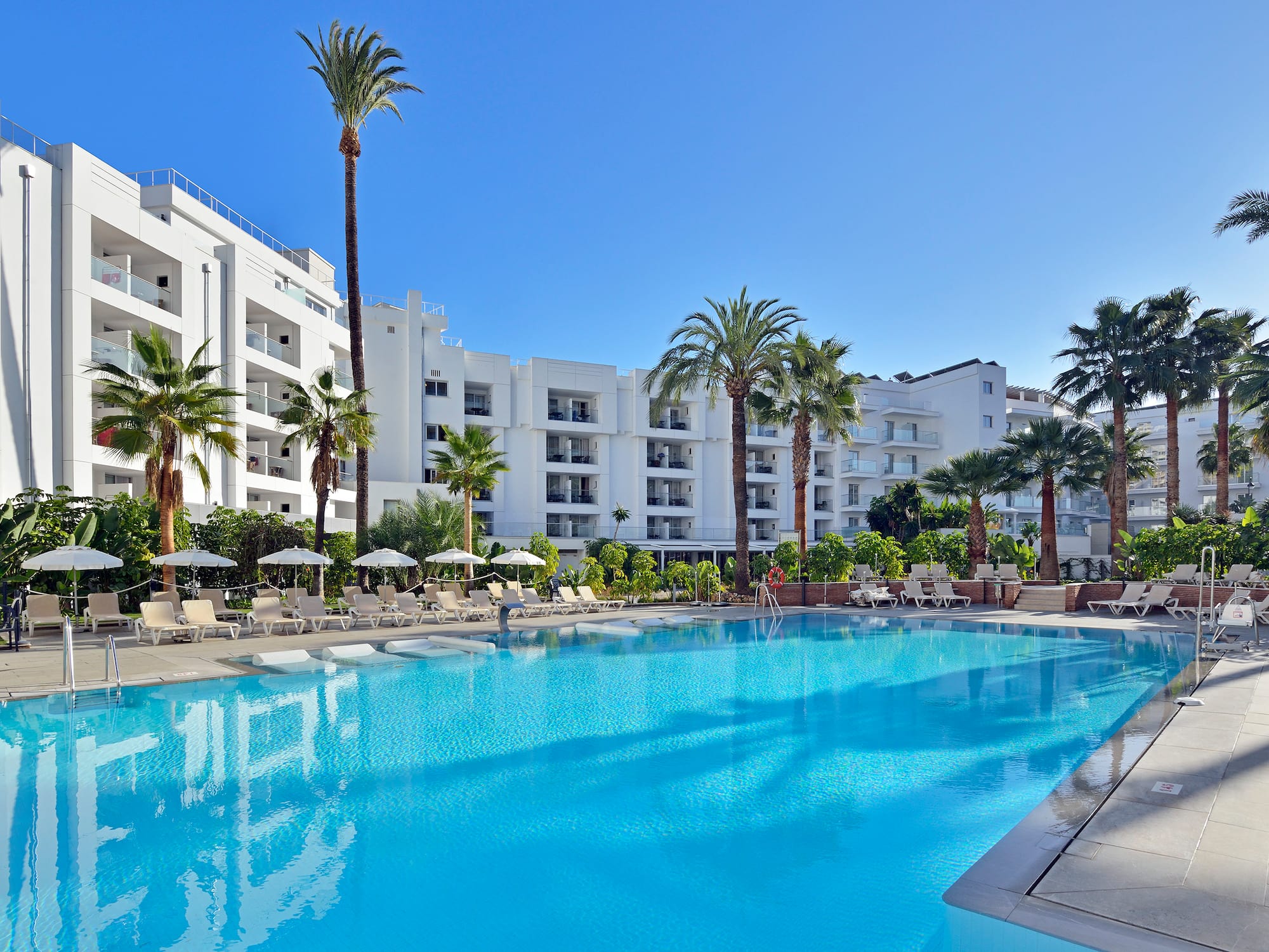 Sunny resort pool with white buildings, palm trees, and lounge chairs.