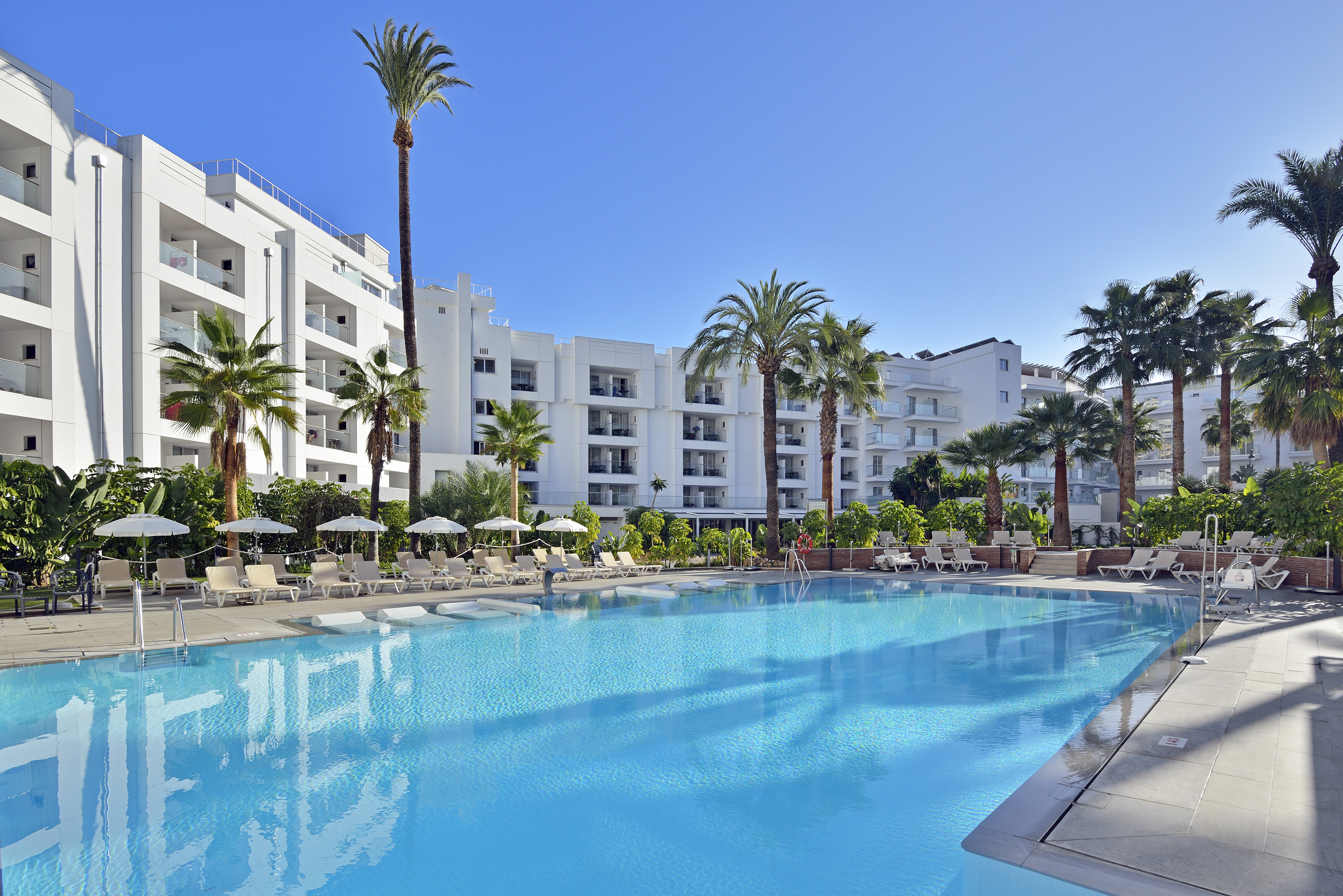 Sunny resort pool with white buildings, palm trees, and lounge chairs.