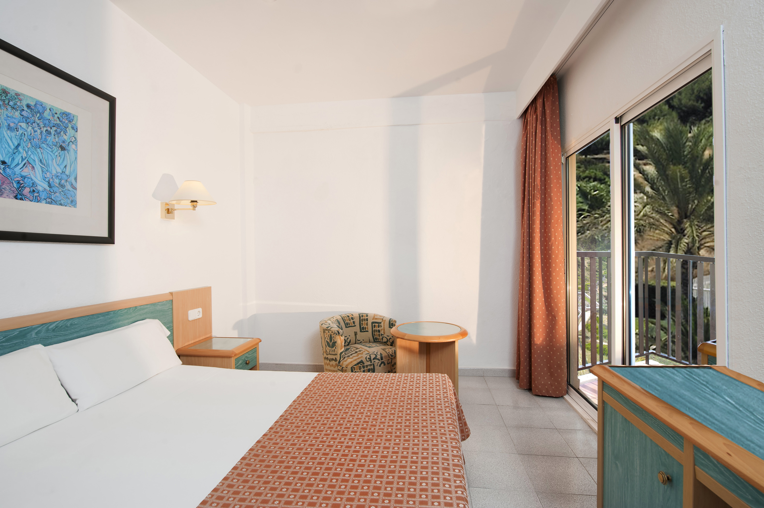 Hotel room with bed, armchair, and balcony overlooking palm trees.