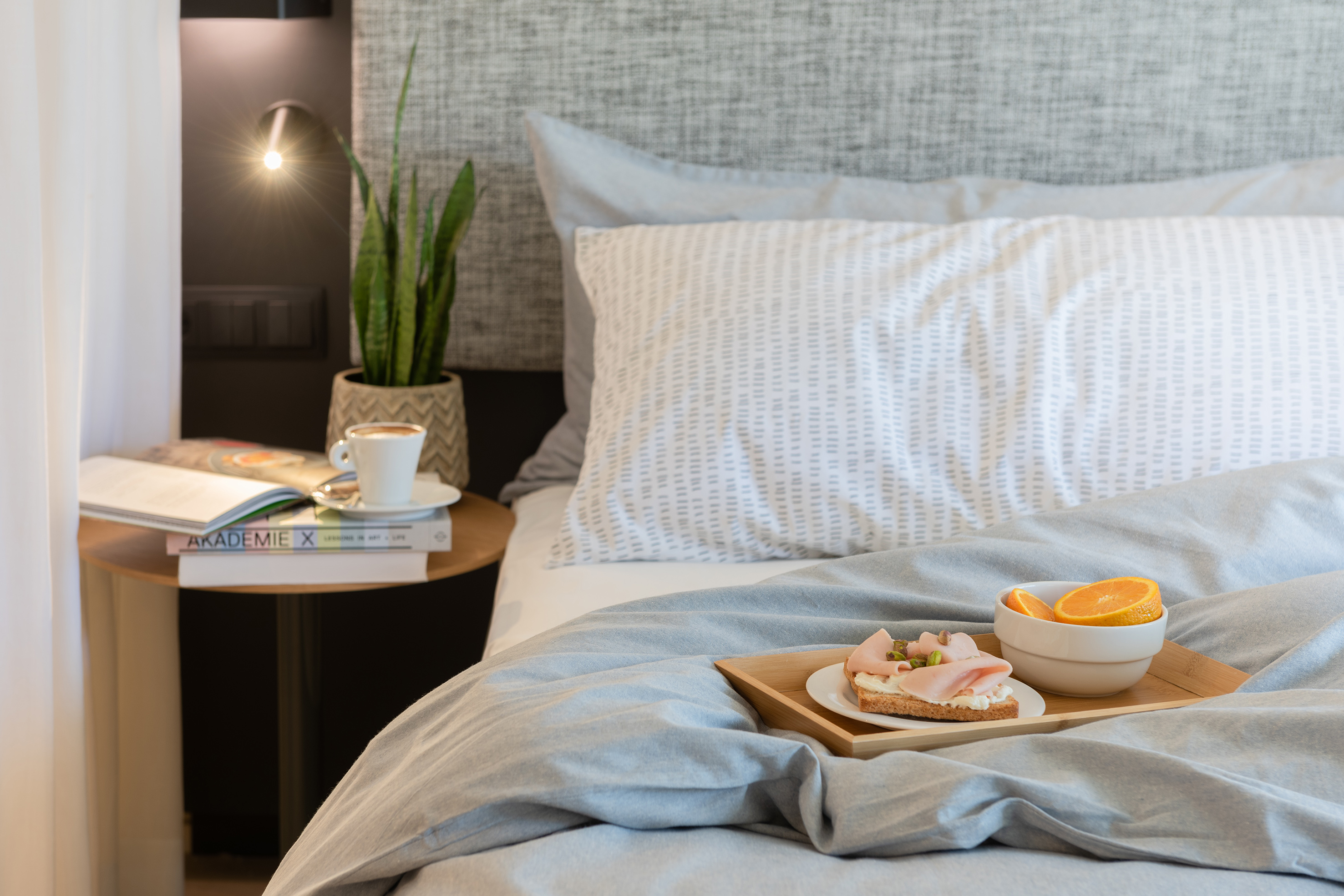 Cozy bedroom with breakfast in bed: sandwich, oranges, coffee, plant, and lamp.