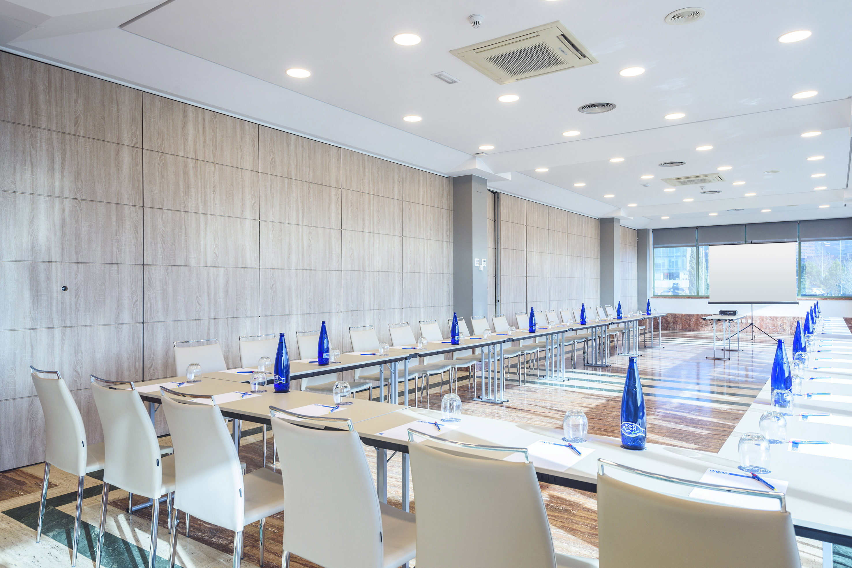 Conference room setup with meeting tables, chairs, water bottles, and screen.