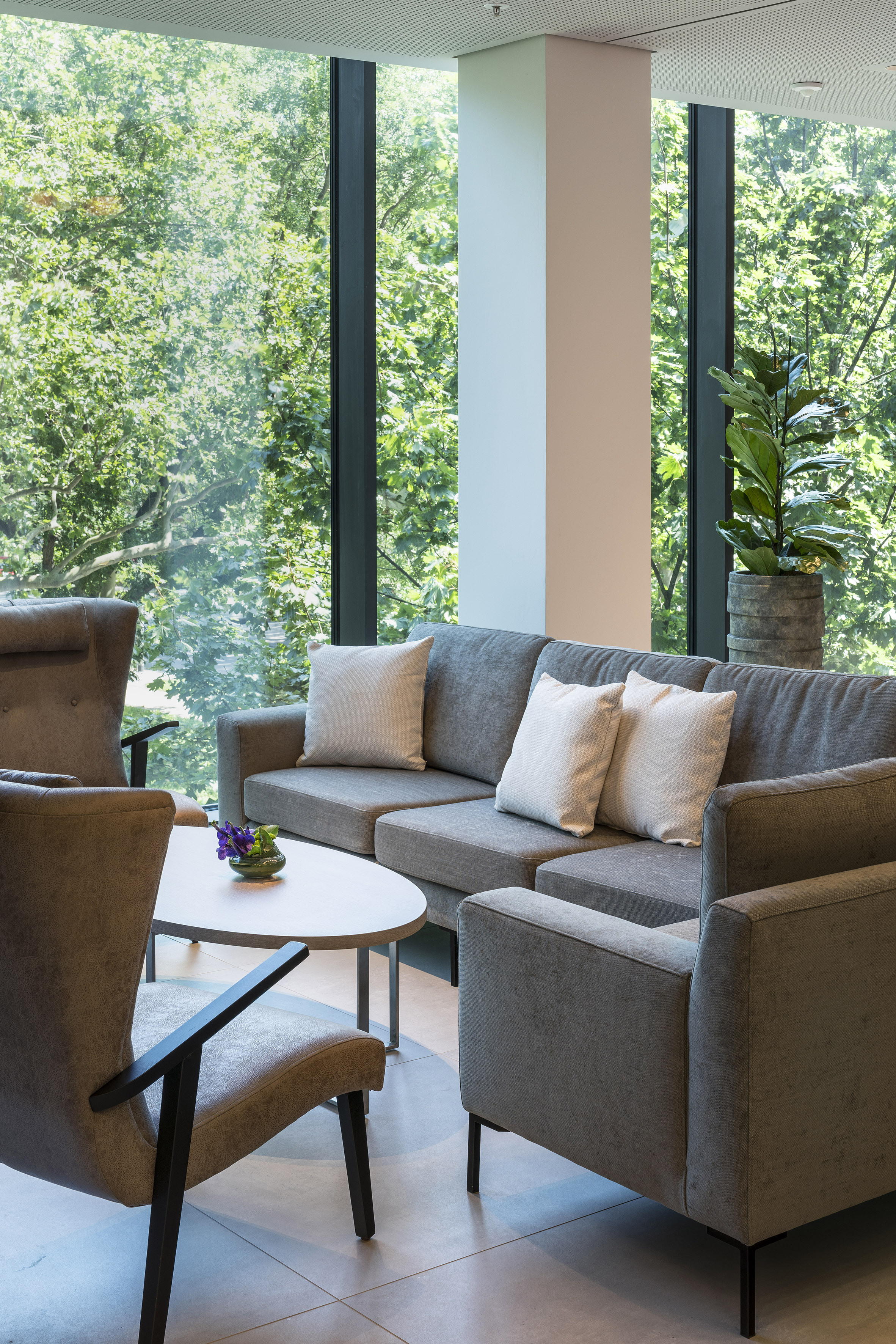 Modern lobby lounge with gray sofa, armchairs, and large window to green trees.