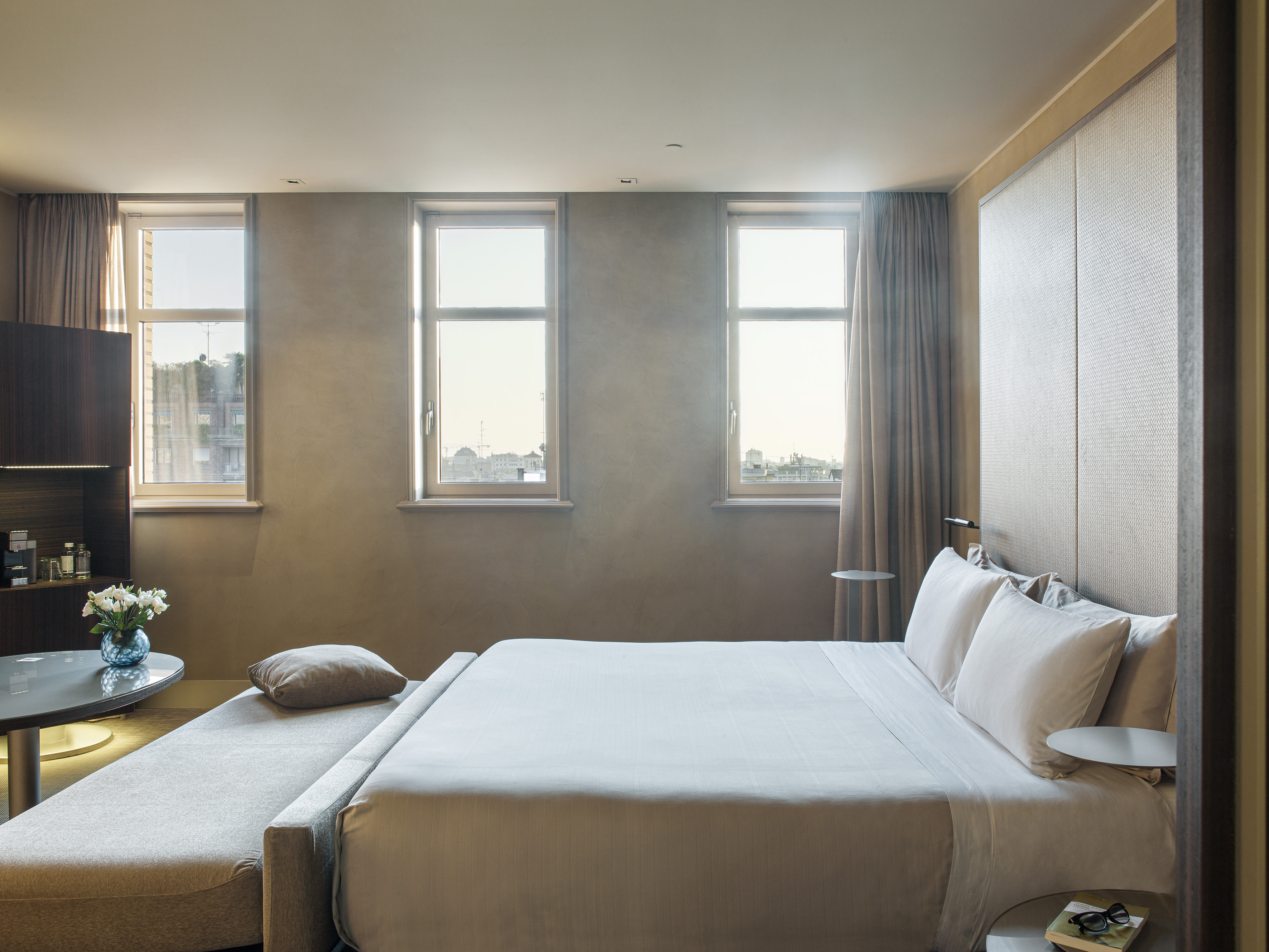 Modern hotel room with large bed, chaise lounge, bright windows, and stylish decor.