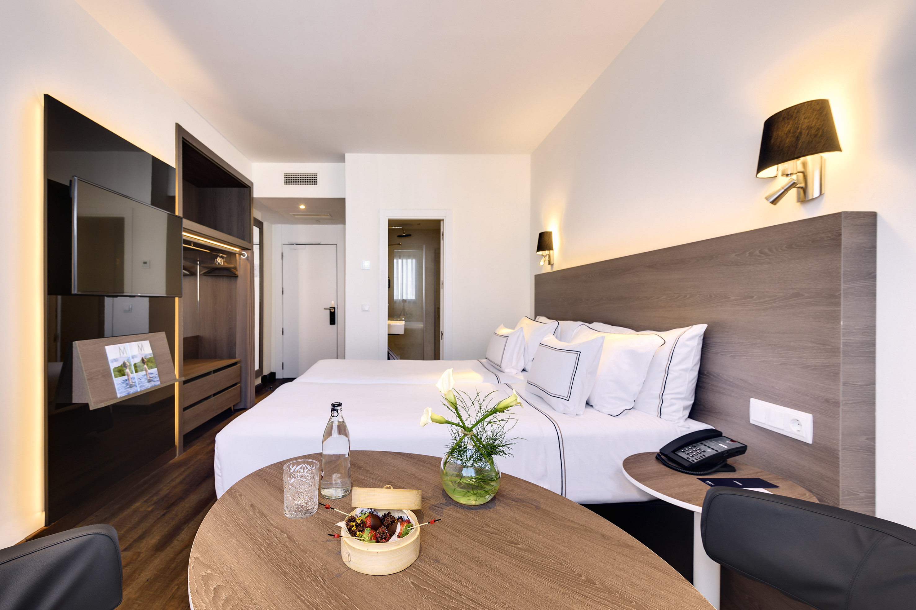 Modern hotel room with twin beds, wood table, TV, and guest amenities.