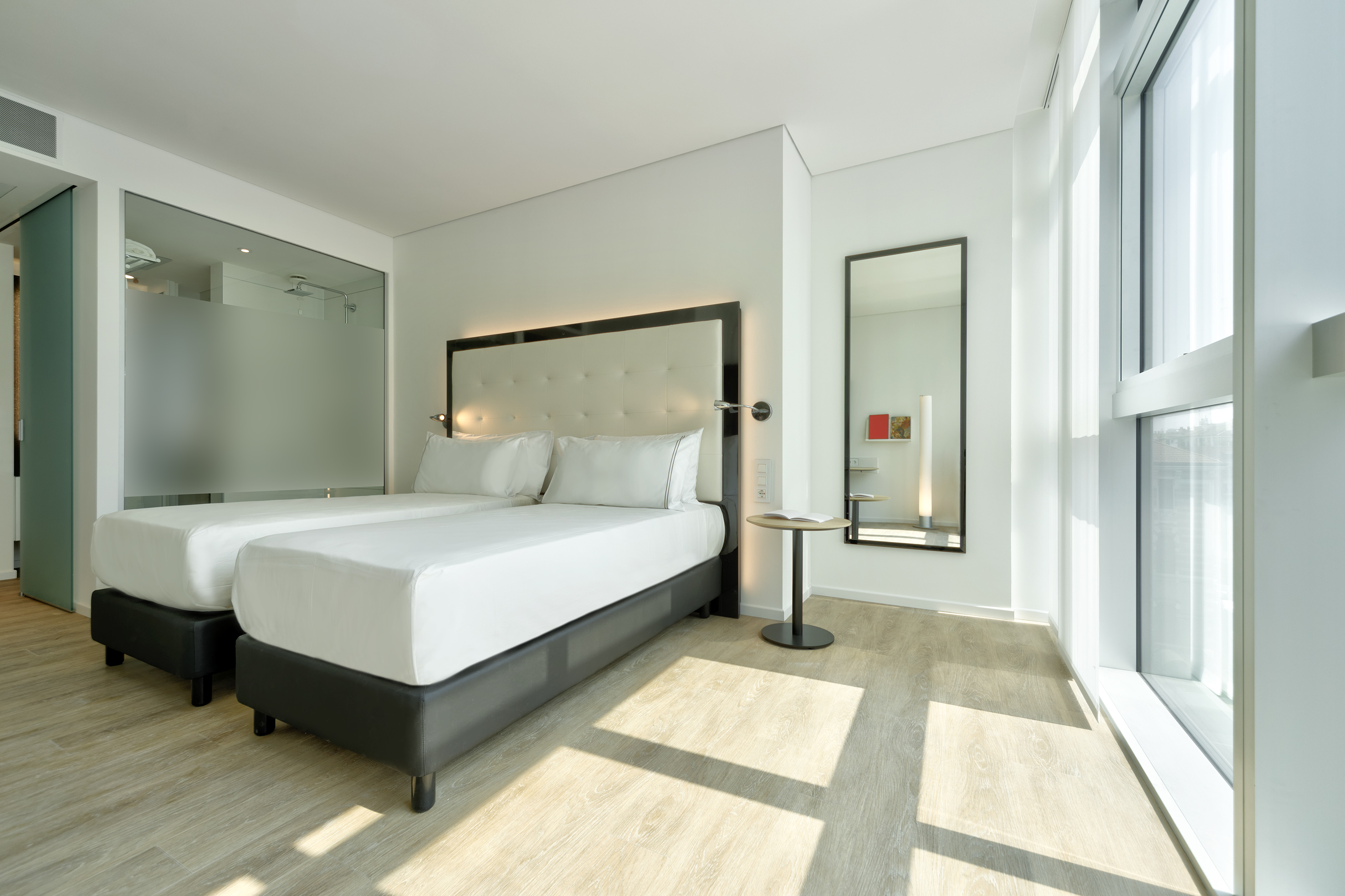 Bright modern hotel room with twin beds, large window, and wood floor.