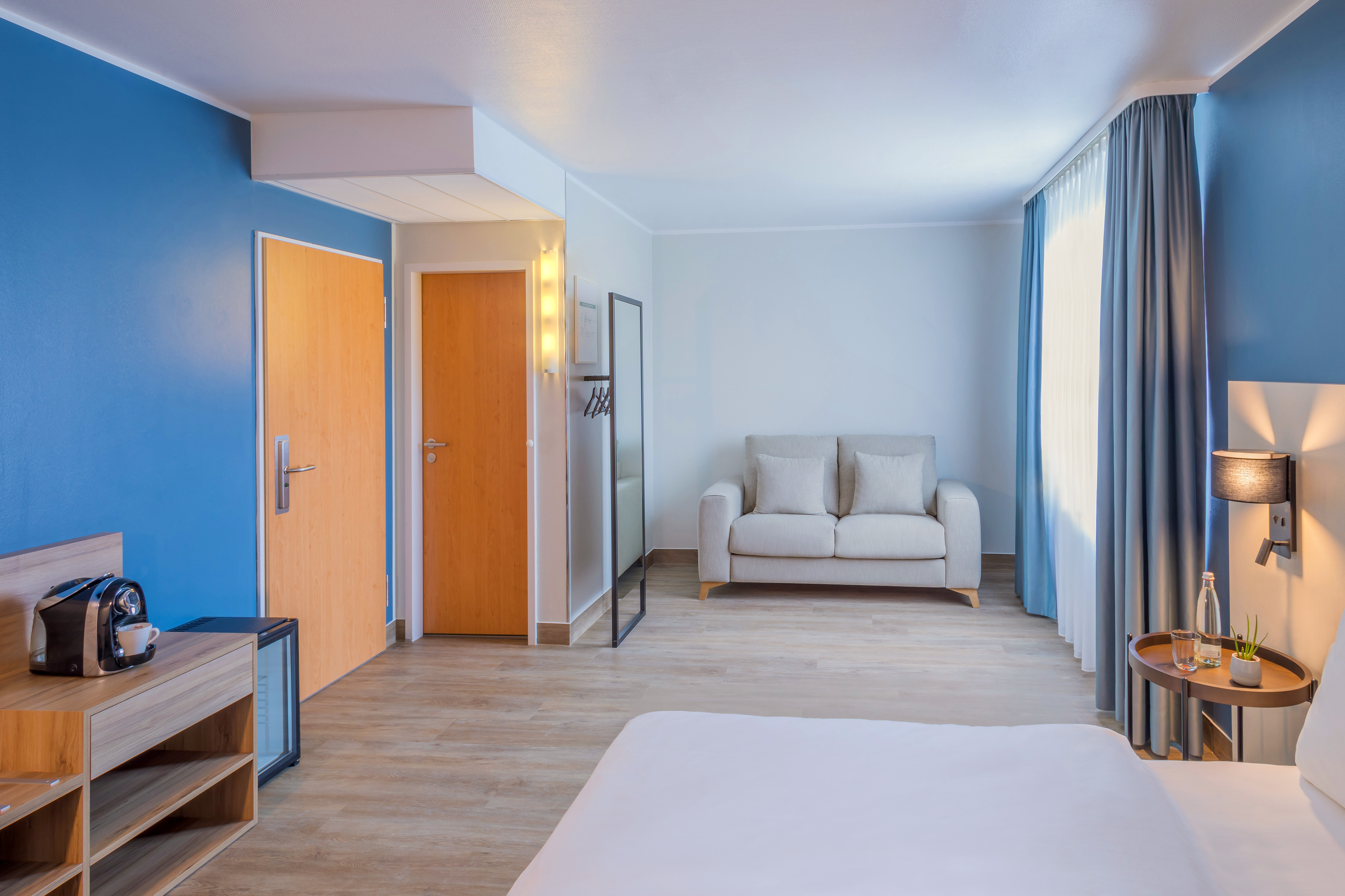 Hotel room with blue accent wall, wood floor, beige sofa, and coffee station.
