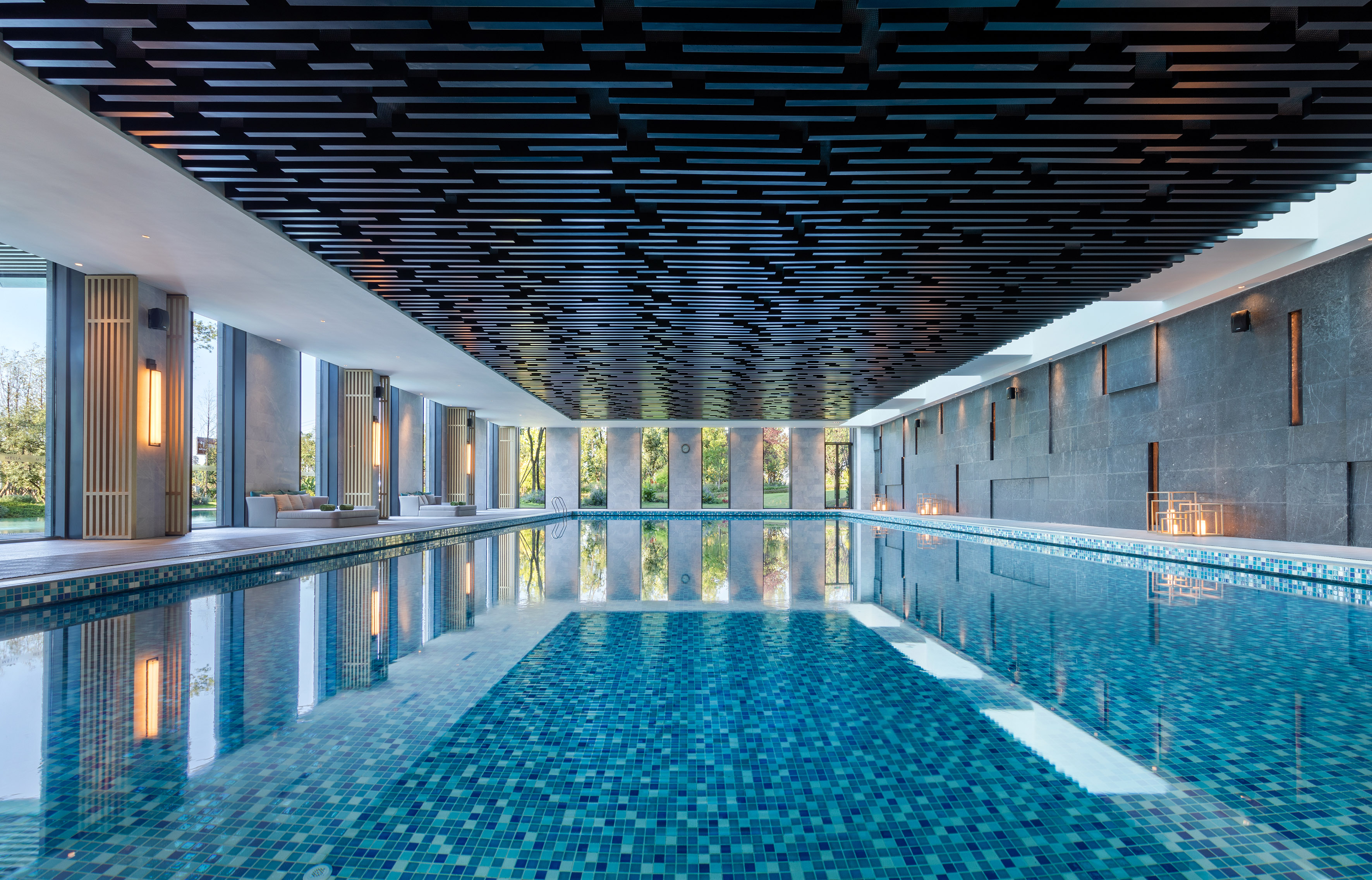 Modern indoor pool with blue tiles, slatted ceiling, and window views.
