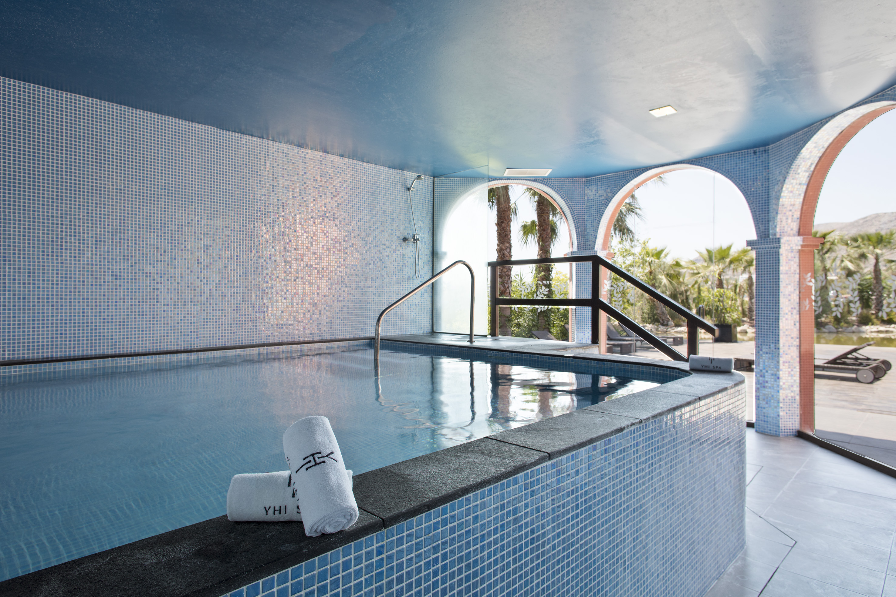 Blue tiled indoor spa pool, arched windows, outdoor palm trees. YHI SPA.