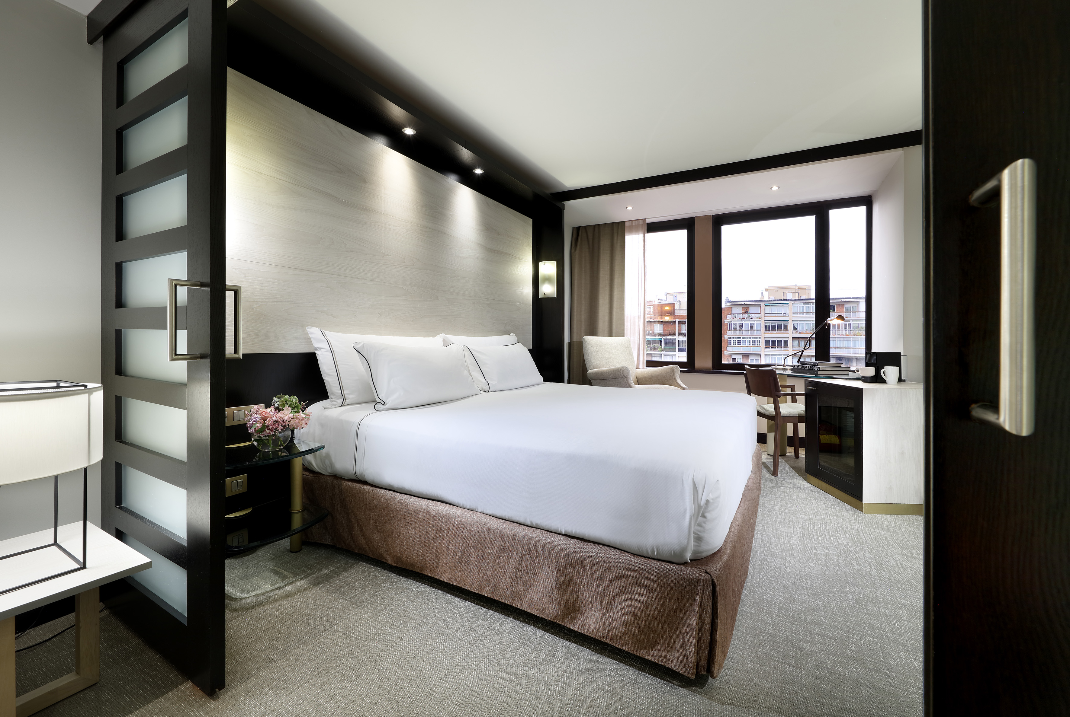 Modern hotel bedroom with white bed, dark wood, desk area, and city view.