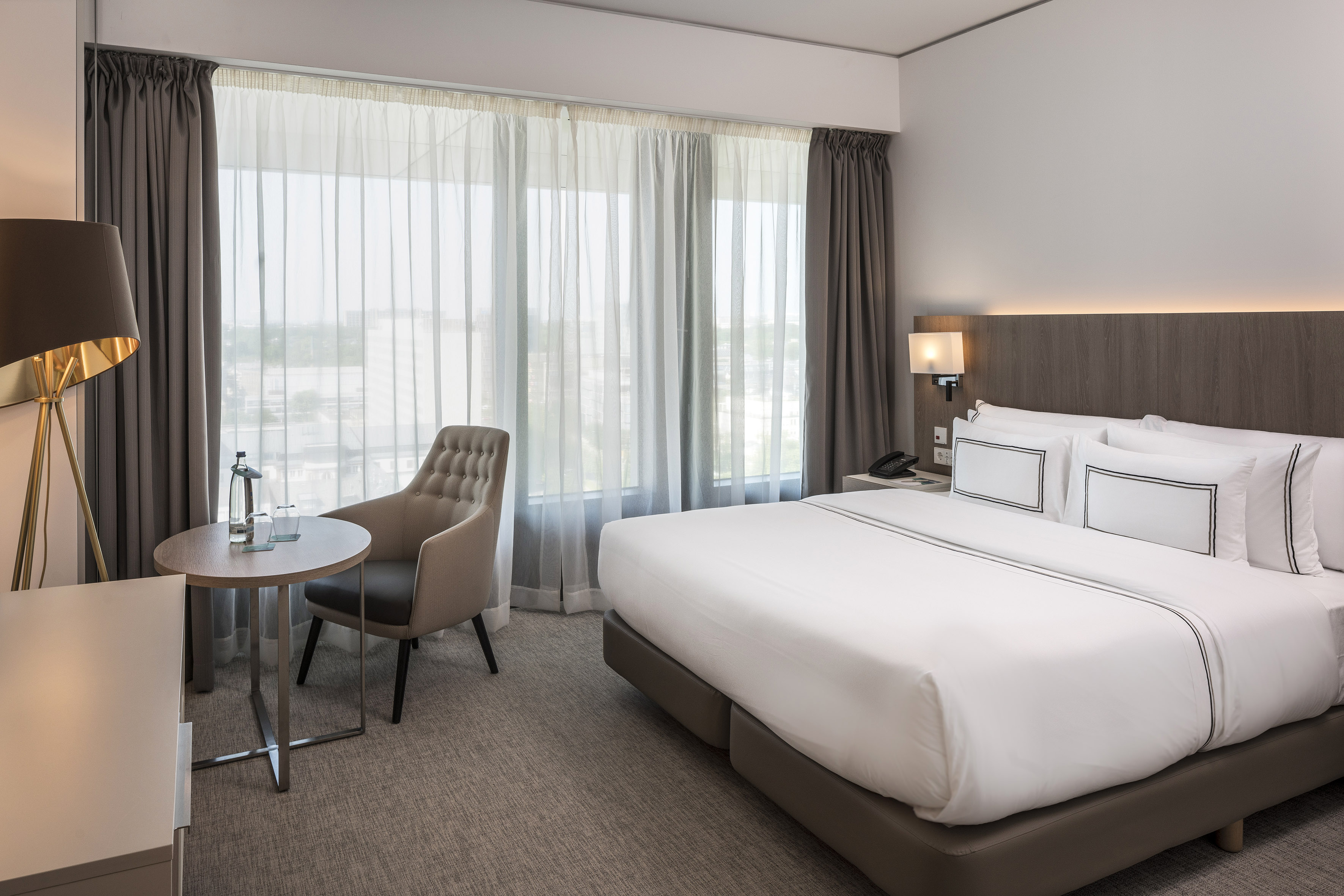Modern hotel bedroom with king bed, armchair, and large window.