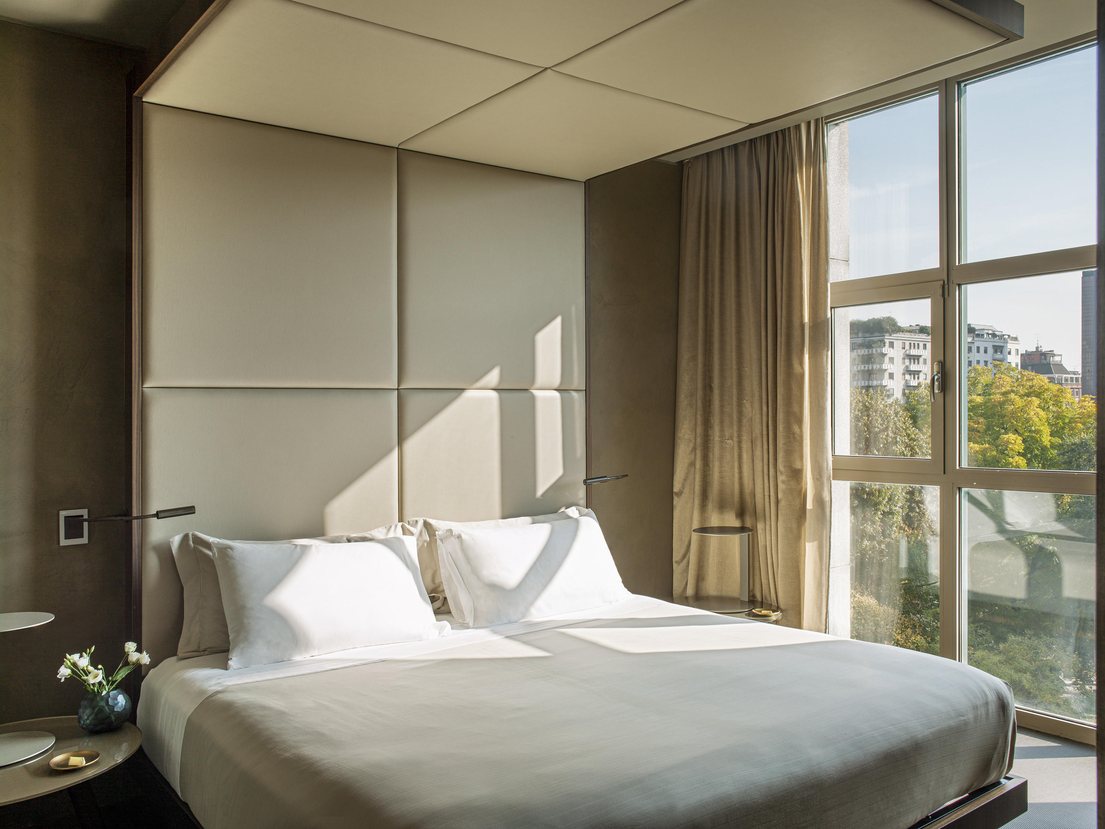 Modern hotel bedroom with neutral decor, padded headboard, and city view window.