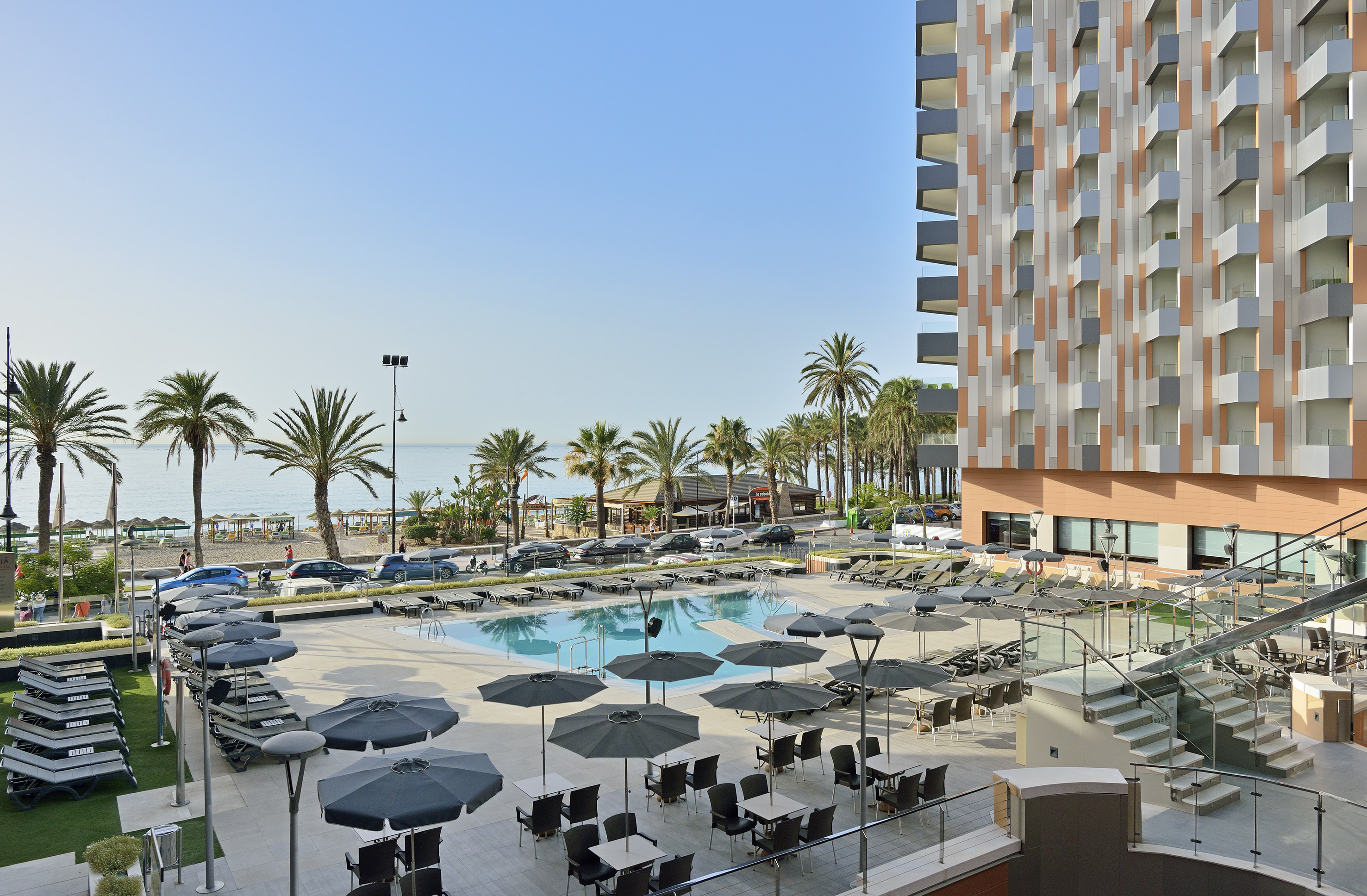 Sunny resort pool, modern hotel, palm trees, and beach overlooking the sea.