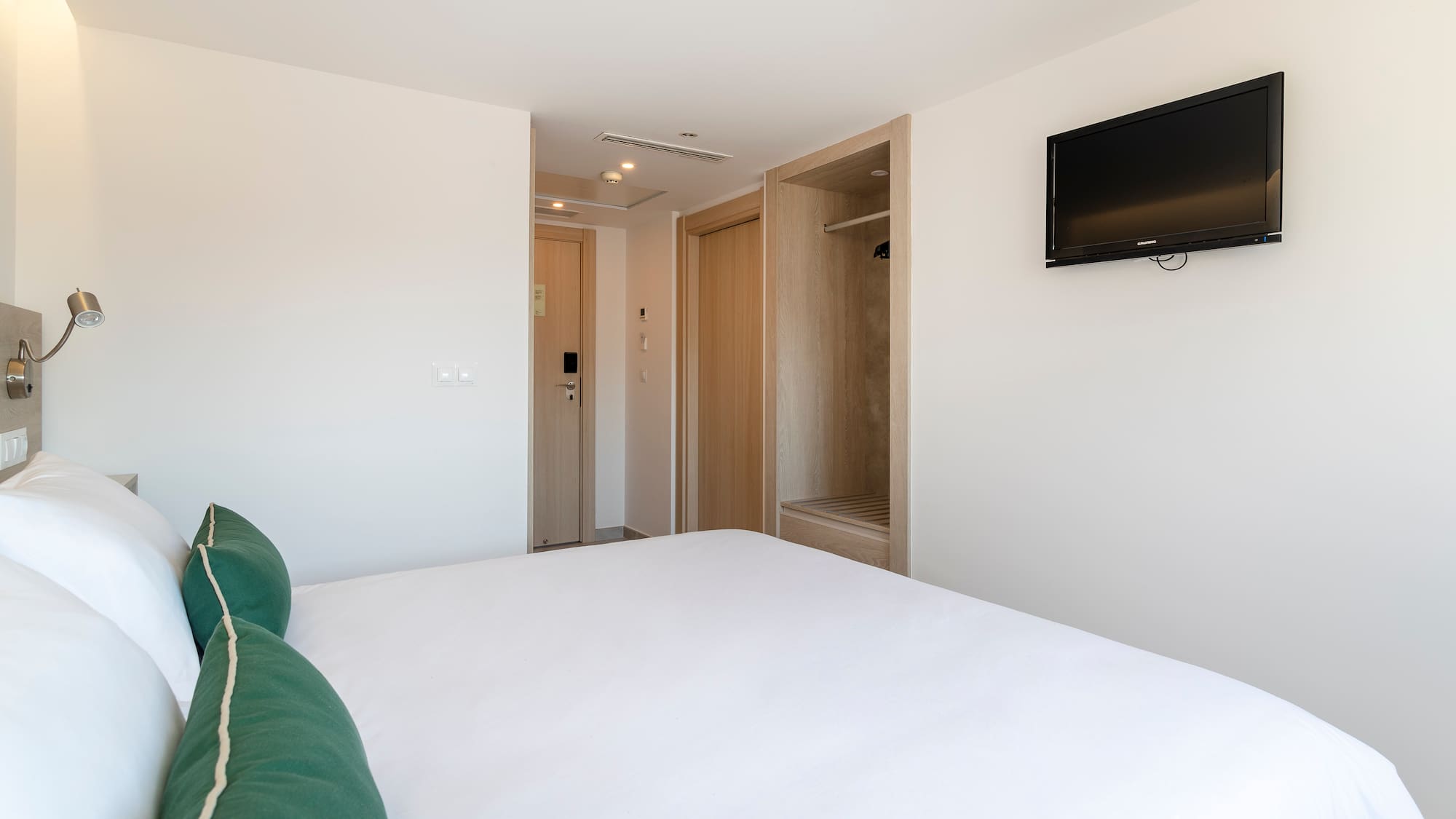 Modern hotel room with white bed, green pillows, wall TV, and closet.